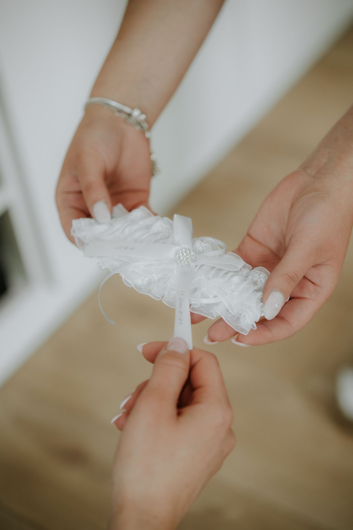 Hands holding a delicate white lace wedding garter with satin ribbon and rhinestone detail.