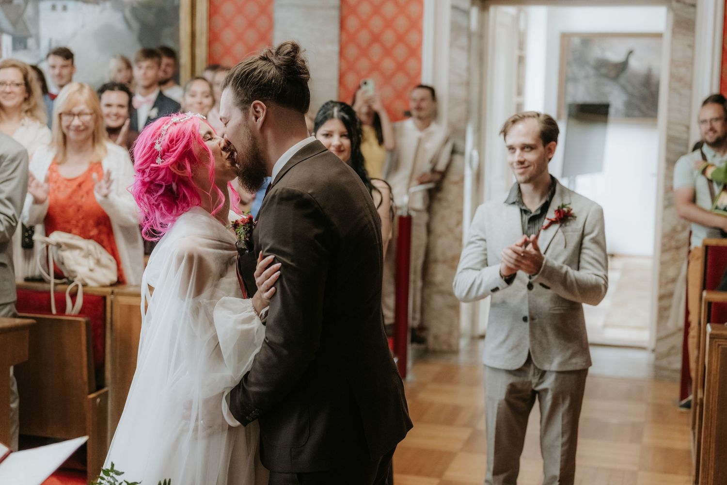 Bride with pink hair and groom share first kiss at church wedding altar while guests applaud in background.