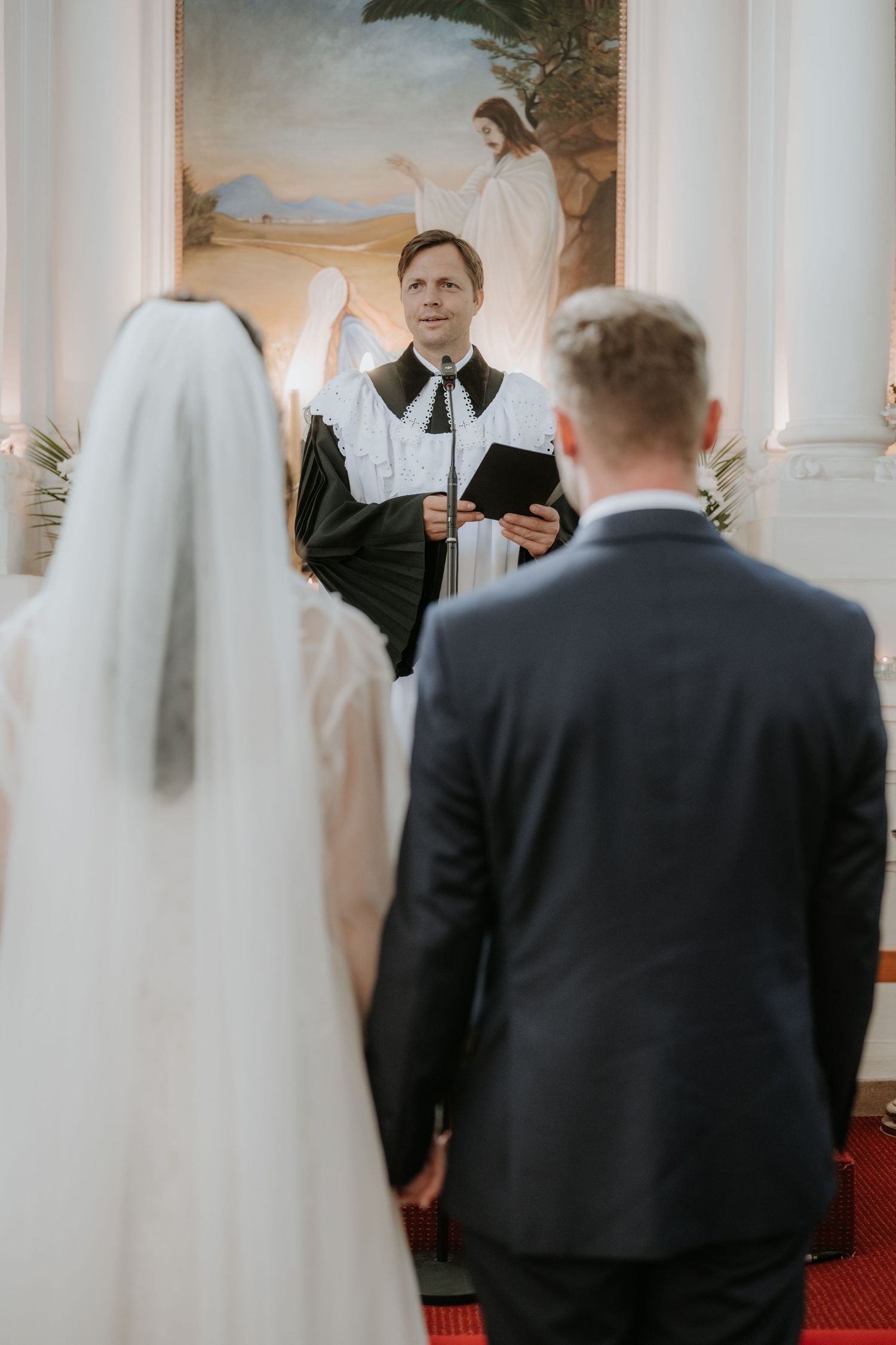 A bride and groom stand before a minister holding a Bible during a church wedding ceremony with religious artwork behind.