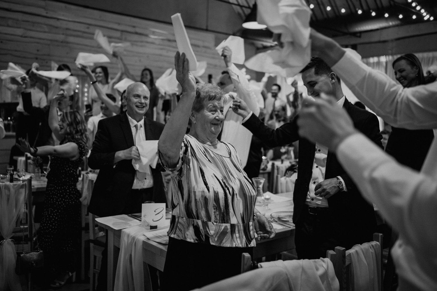 Joyful wedding guests waving white napkins and dancing at a lively indoor reception celebration.