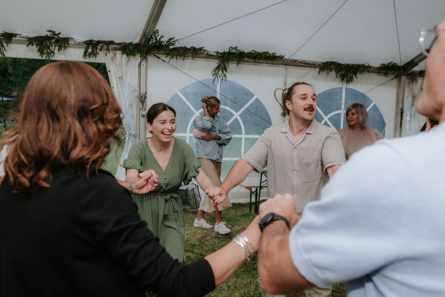 Guests joyfully holding hands in a circle dance under a white tent at an outdoor wedding celebration.