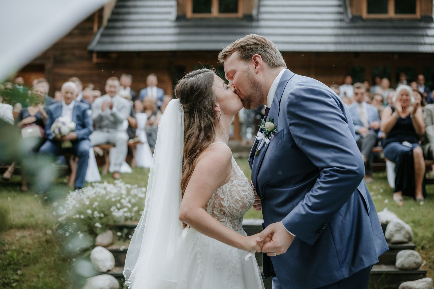 Bride and groom share first kiss at outdoor wedding ceremony with guests seated behind them.