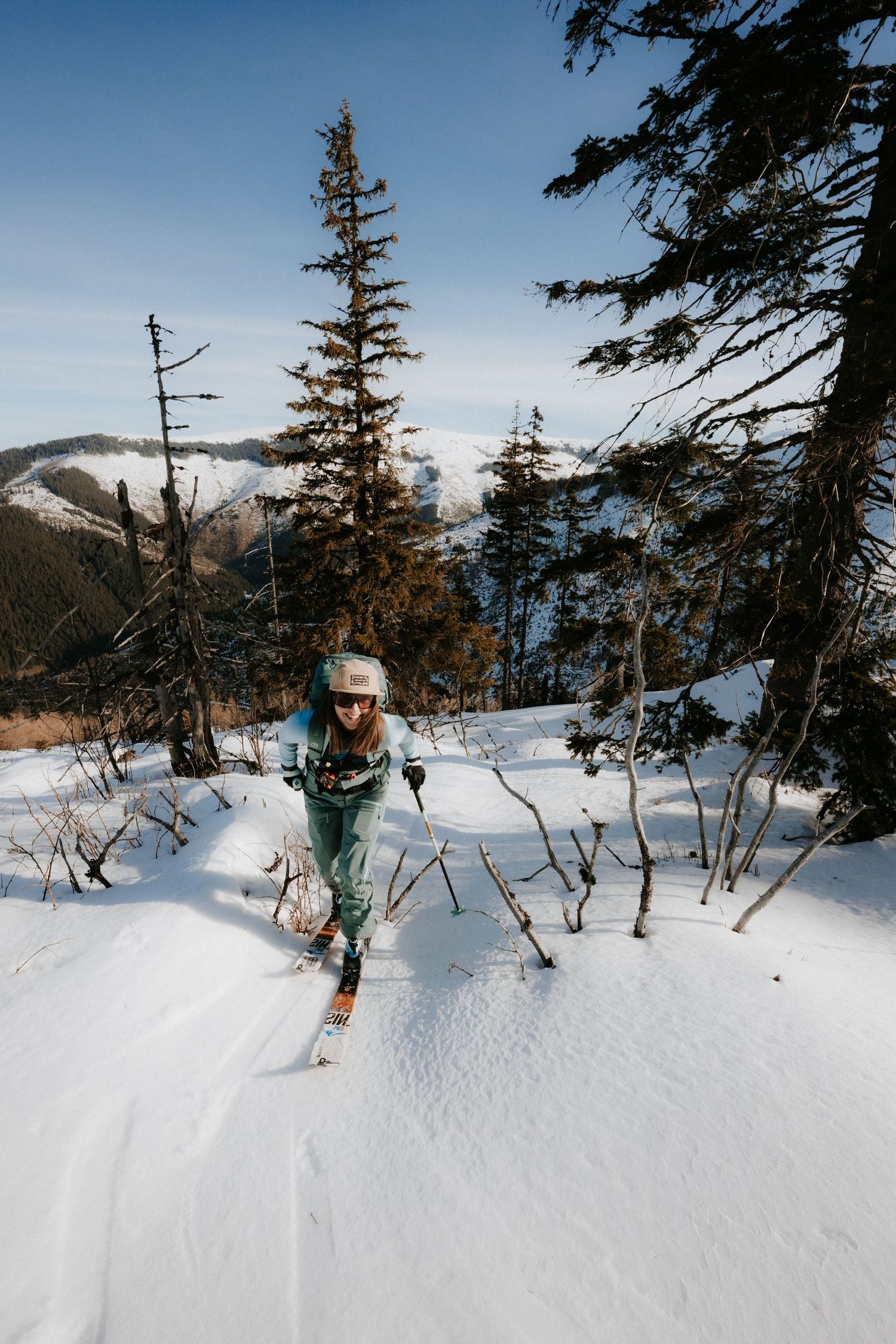Woman in teal ski suit climbing steep snowy mountain slope through sparse pine trees.
