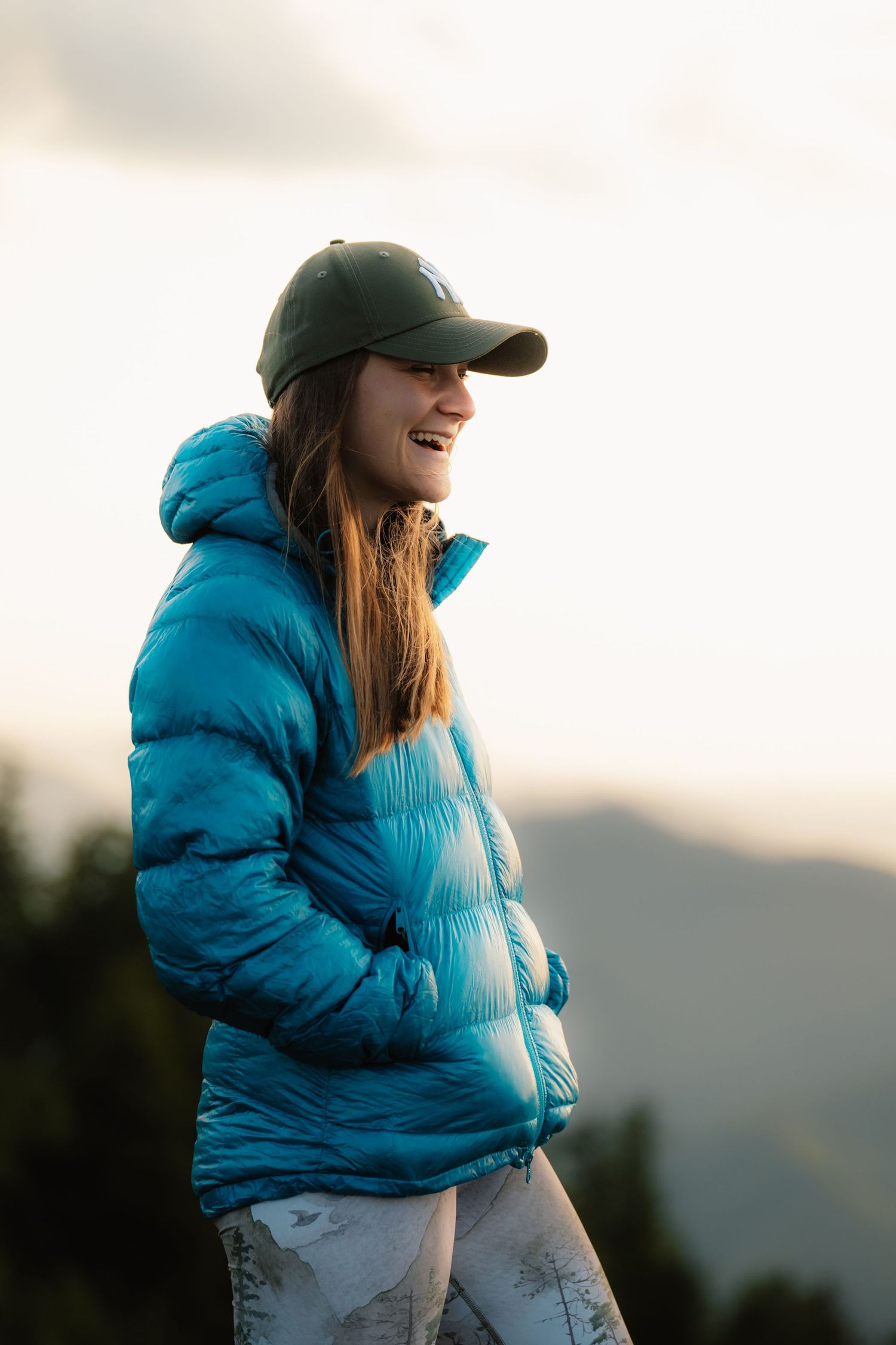 A smiling woman in a teal puffer jacket and green cap stands outdoors with mountain scenery at sunset.