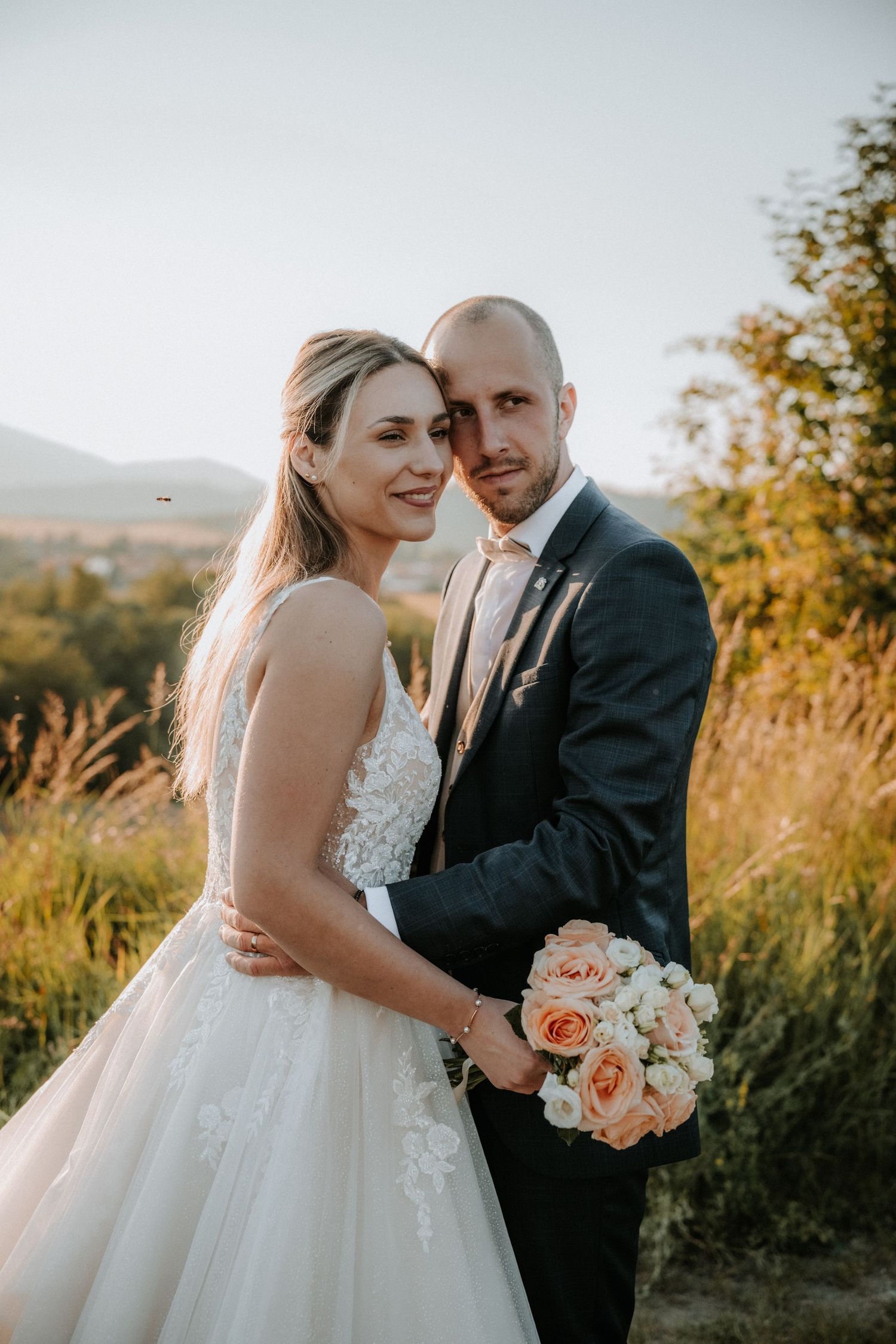 Bride and groom pose together at golden hour, she holds a peach rose bouquet in a lace wedding gown.