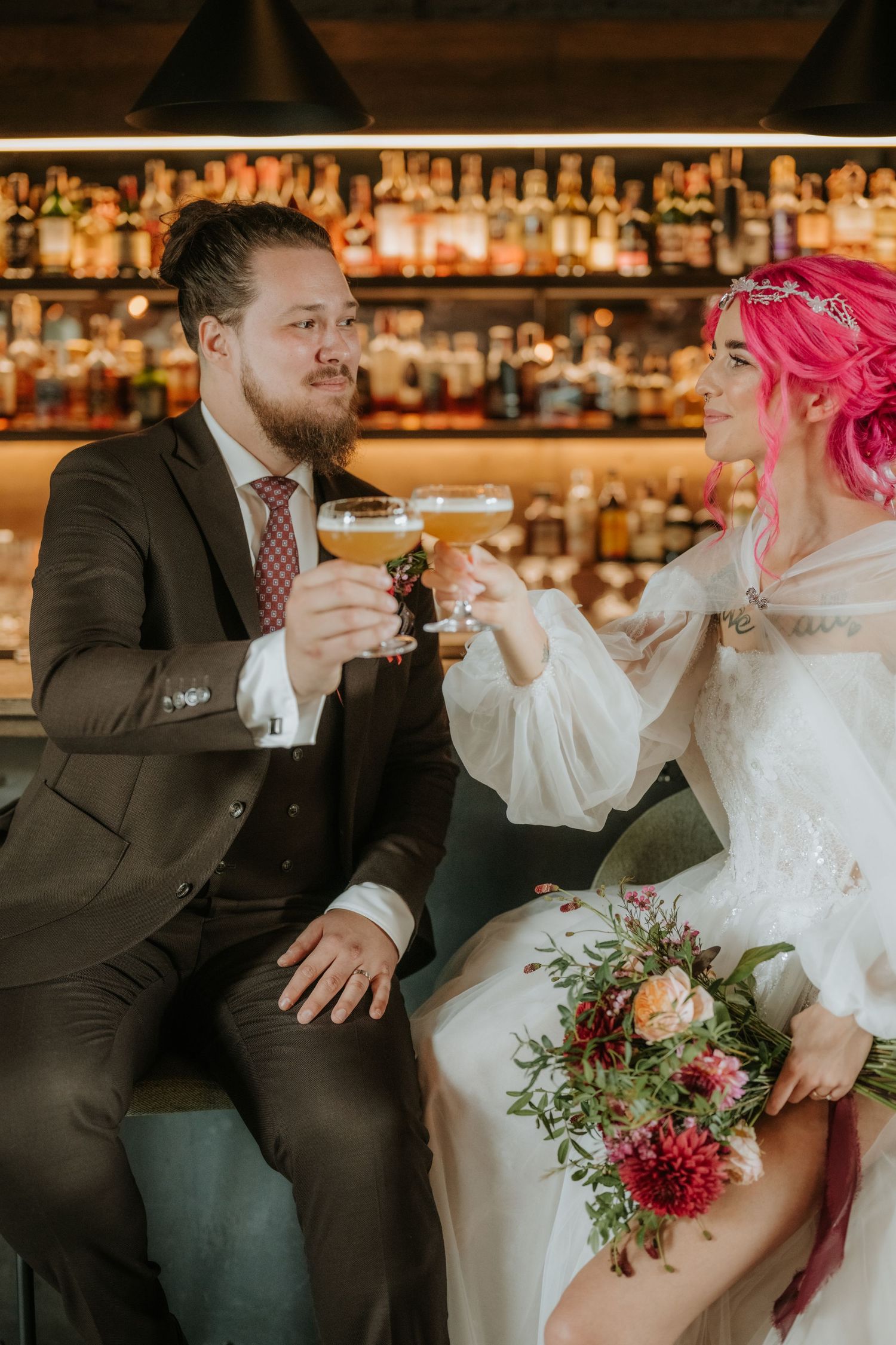 Bride with pink hair and groom in brown suit toasting drinks at a bar with floral bouquet.