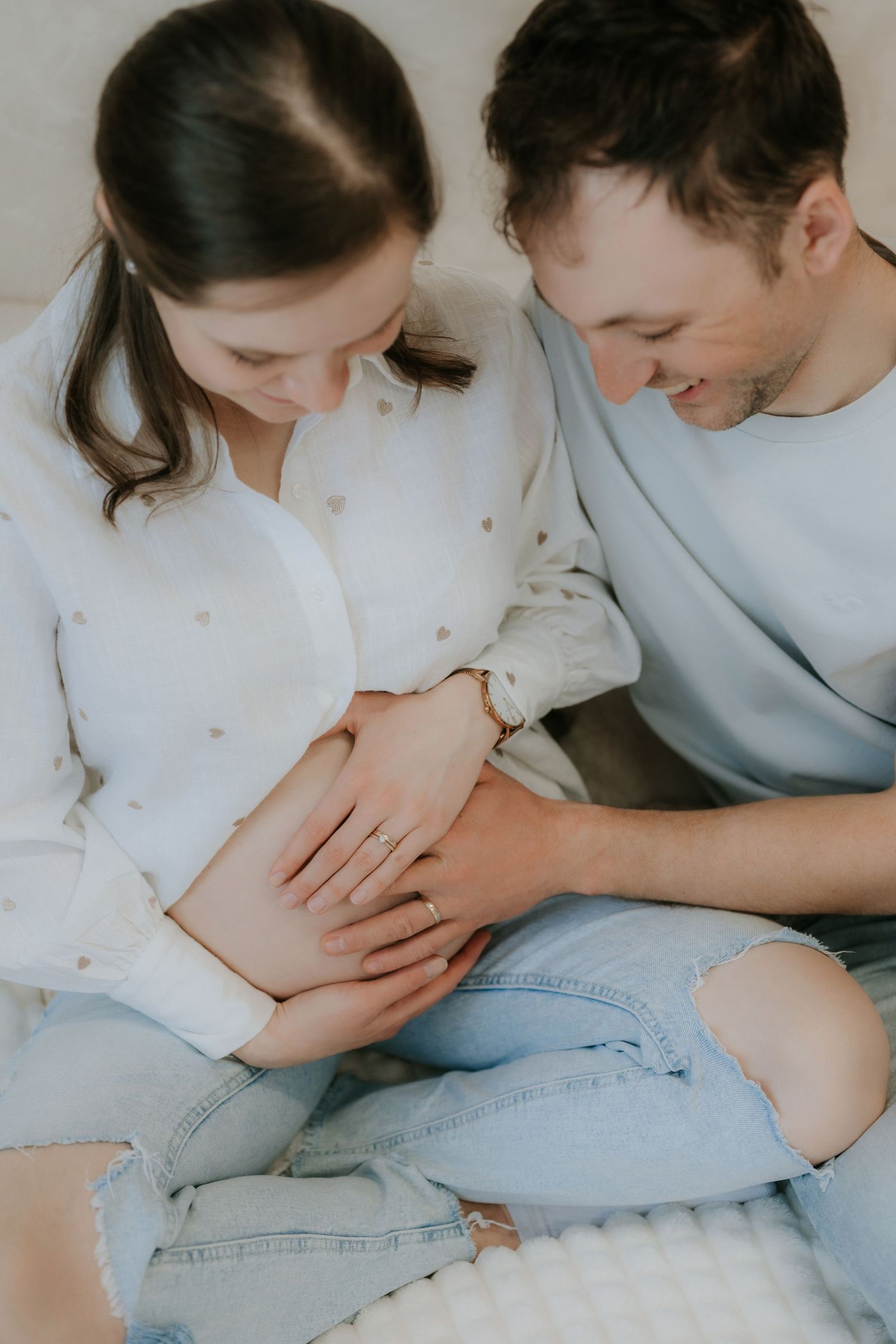 Tender moment of expectant couple sitting together, both looking down at pregnant belly with joy.