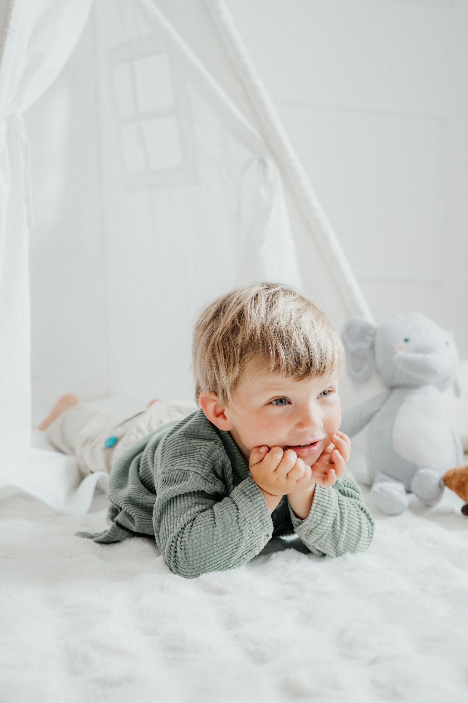 A young blond boy lying on a white bed with his chin resting on his hands near a play tent.