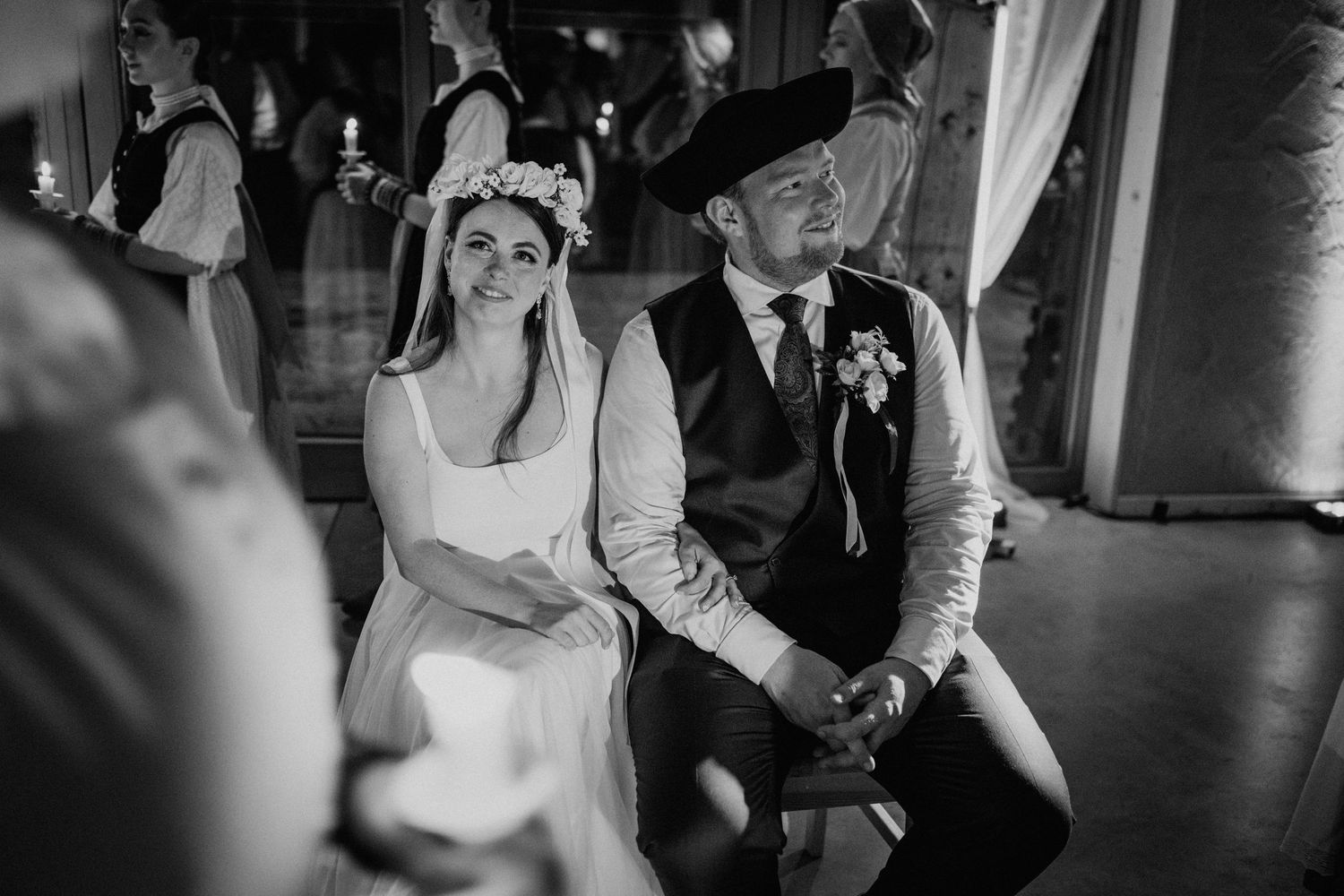 Black and white wedding photo of bride in floral crown and groom in vest sitting together during ceremony.