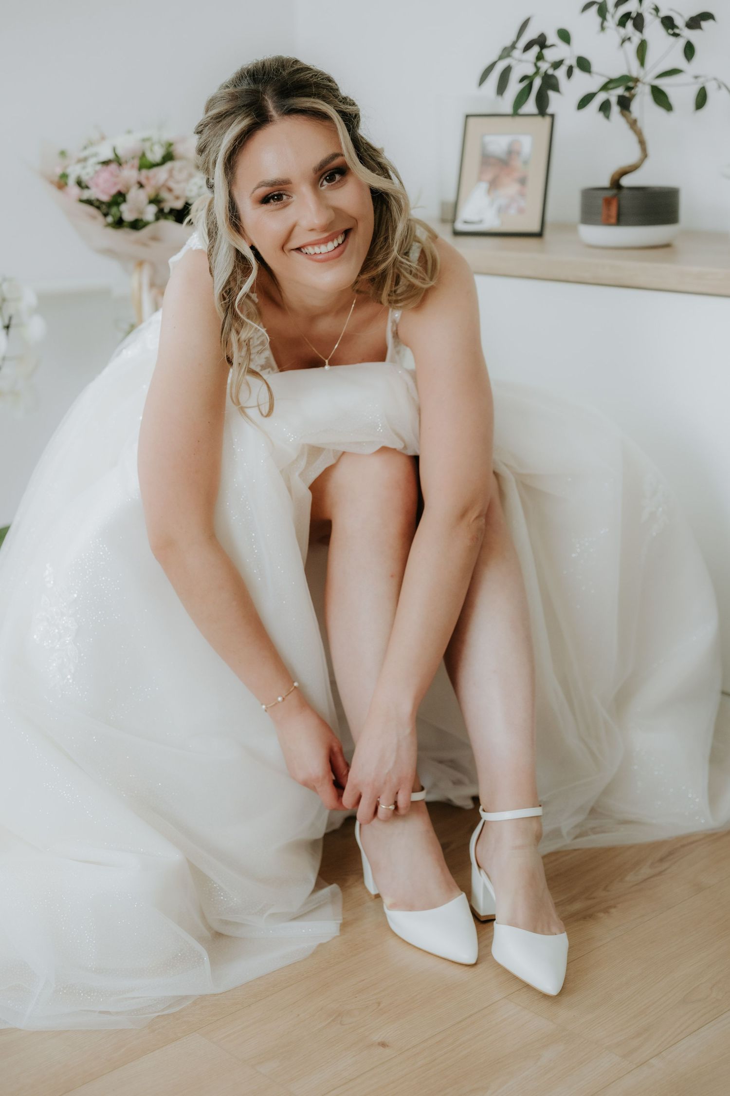 A smiling bride in a white gown fastens her white heeled shoes while seated, with flowers and decor in the background.
