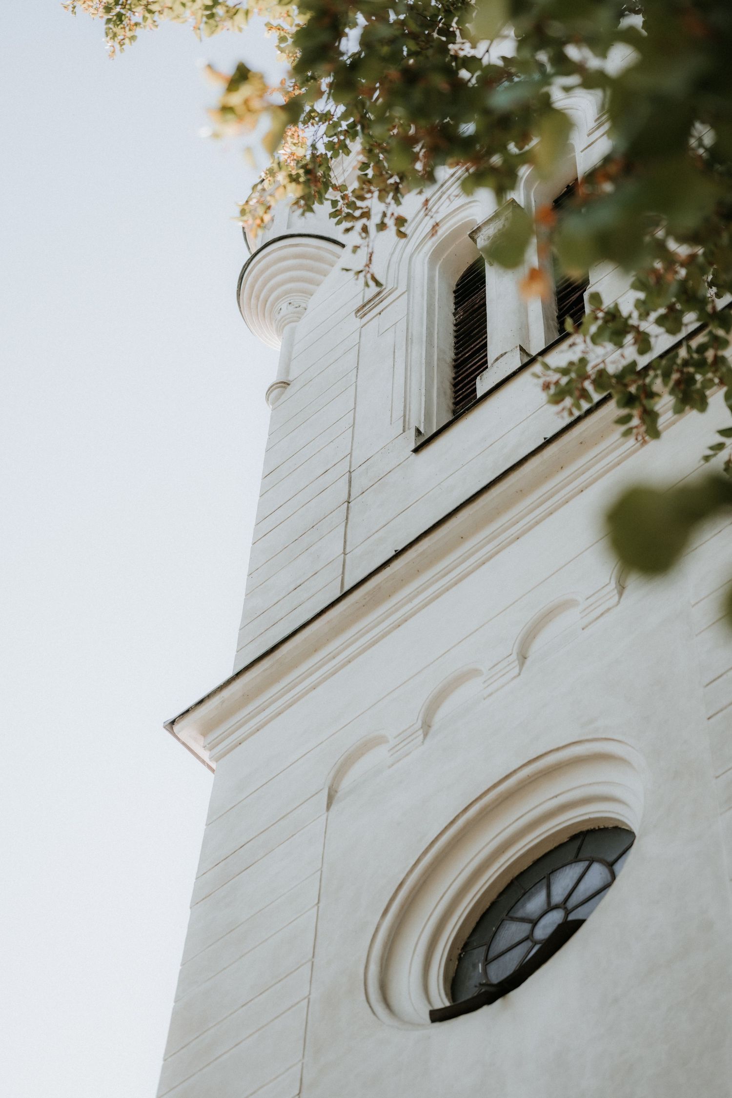 White church tower with ornate round window and bell tower, framed by green tree branches against a pale sky.