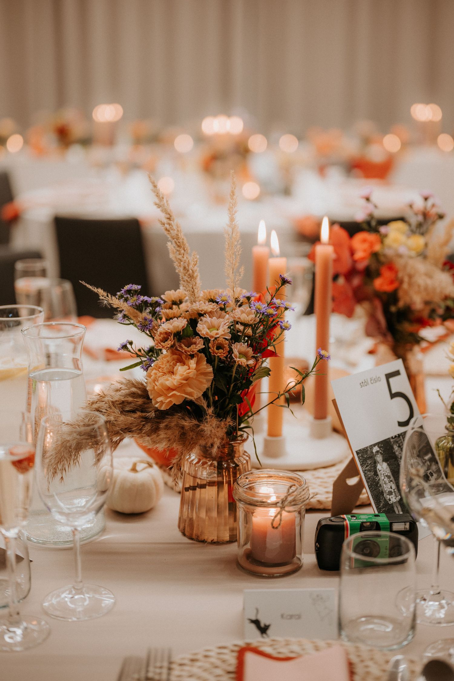 Elegant fall wedding reception table with orange candles, pampas grass floral centerpiece, and table number 5 on white linen.