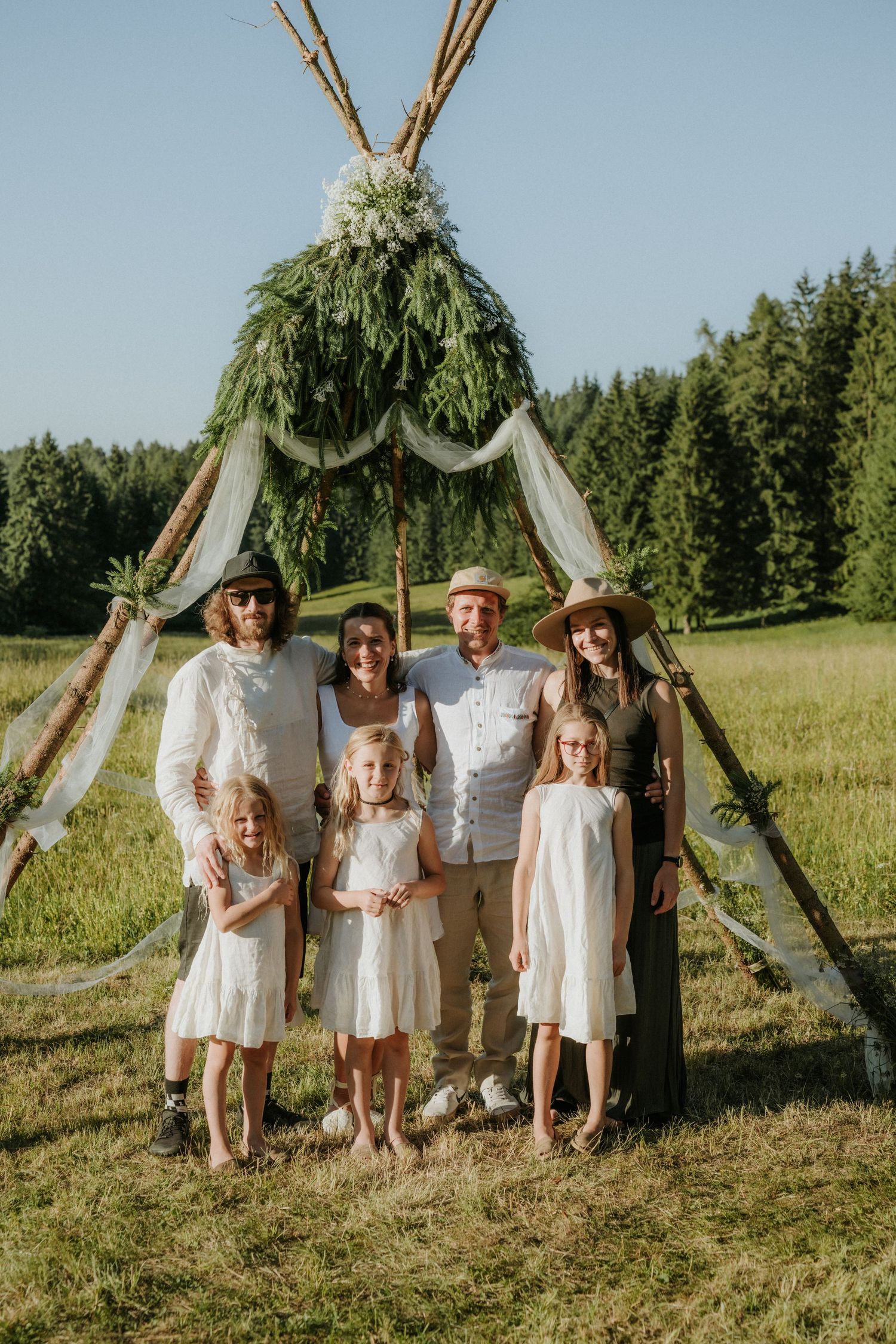Family posing in front of a boho teepee arch with greenery at an outdoor wedding ceremony in a meadow.