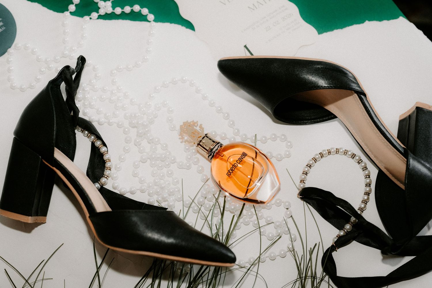Black block-heel pumps with pearl ankle straps and an amber perfume bottle on a white floral surface.