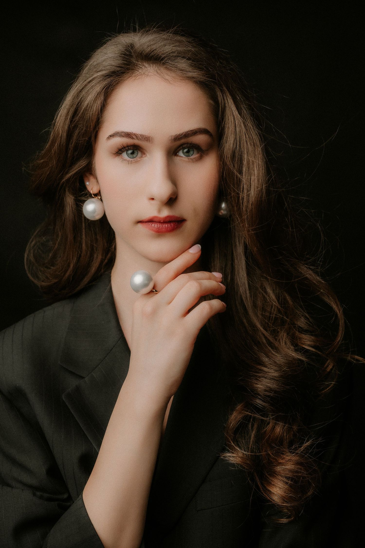 Elegant brunette woman with pearl earrings and ring in dark blazer on black background.