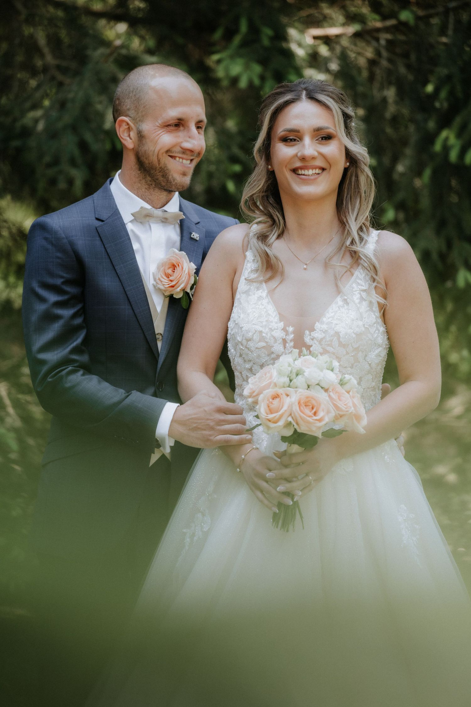 Smiling bride and groom pose outdoors, she holds peach roses bouquet, he wears navy suit with matching boutonniere.
