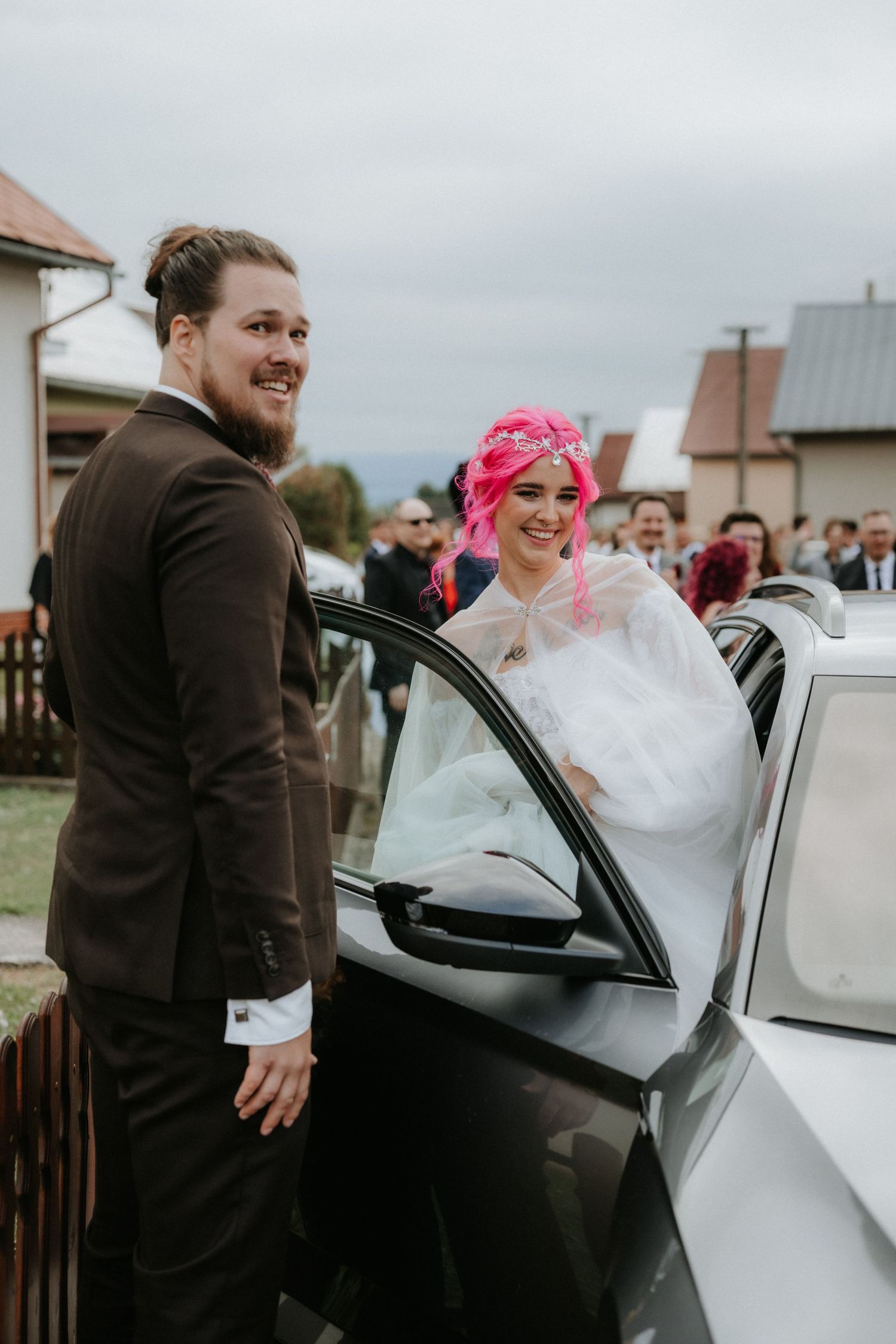 Bride with pink hair in white dress and groom in brown suit smiling beside a black car at a wedding.
