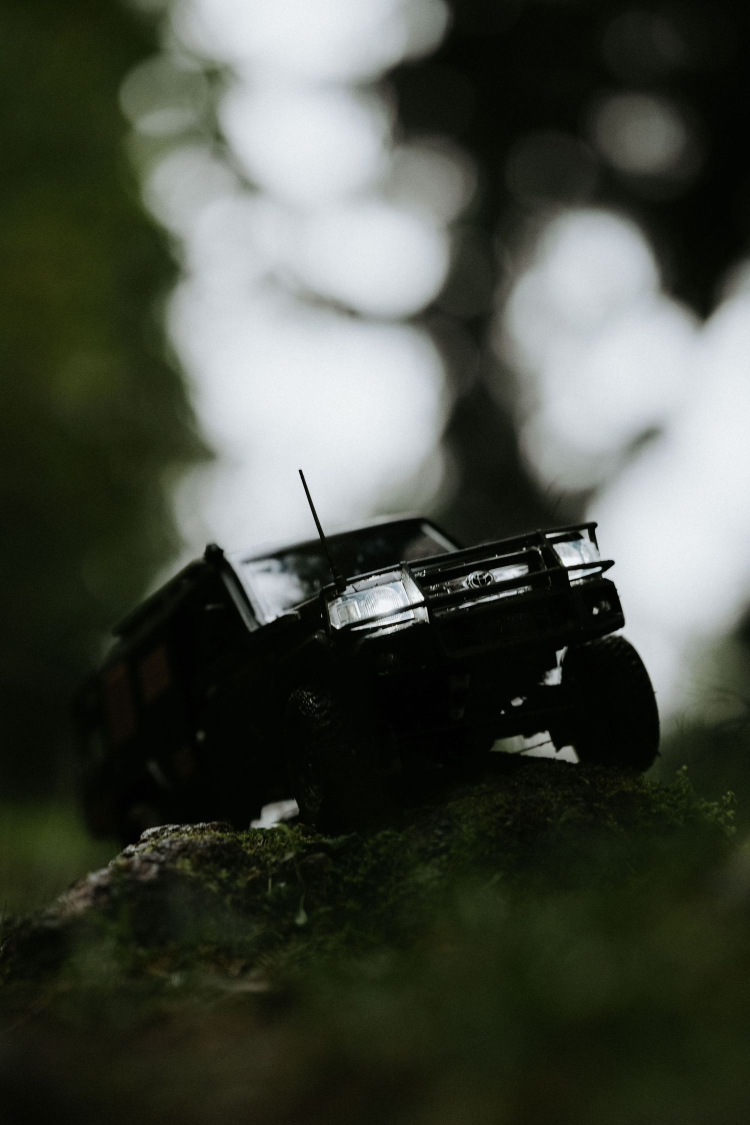 A dark toy off-road RC car climbing a mossy rock in a forest setting with bokeh background.