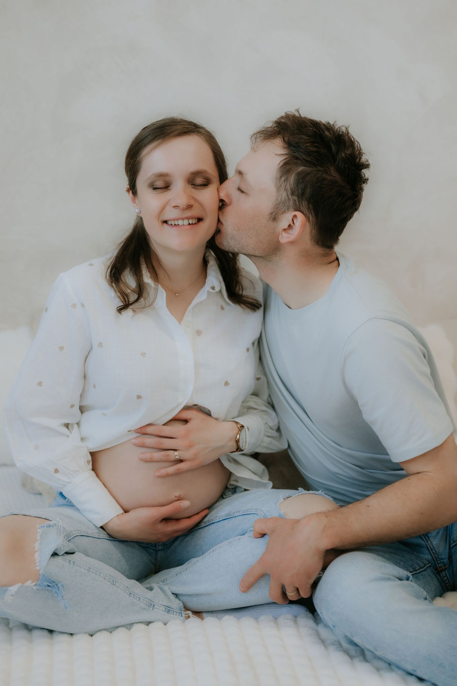 Loving partner kissing smiling pregnant woman on cheek while sitting together during maternity session.
