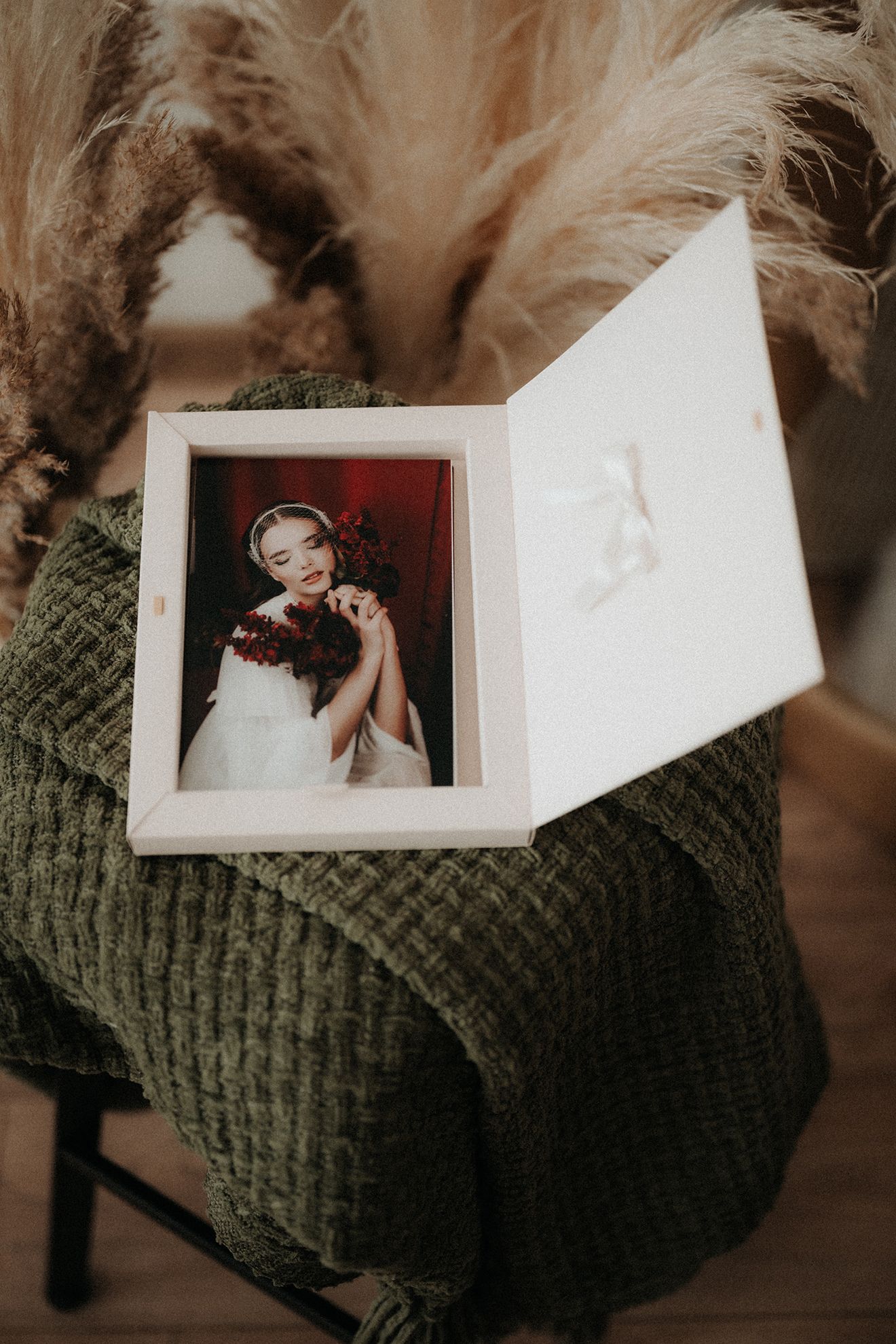 Open white keepsake box with bridal portrait photo on green textured blanket with pampas grass backdrop. Liptovský Mikuláš