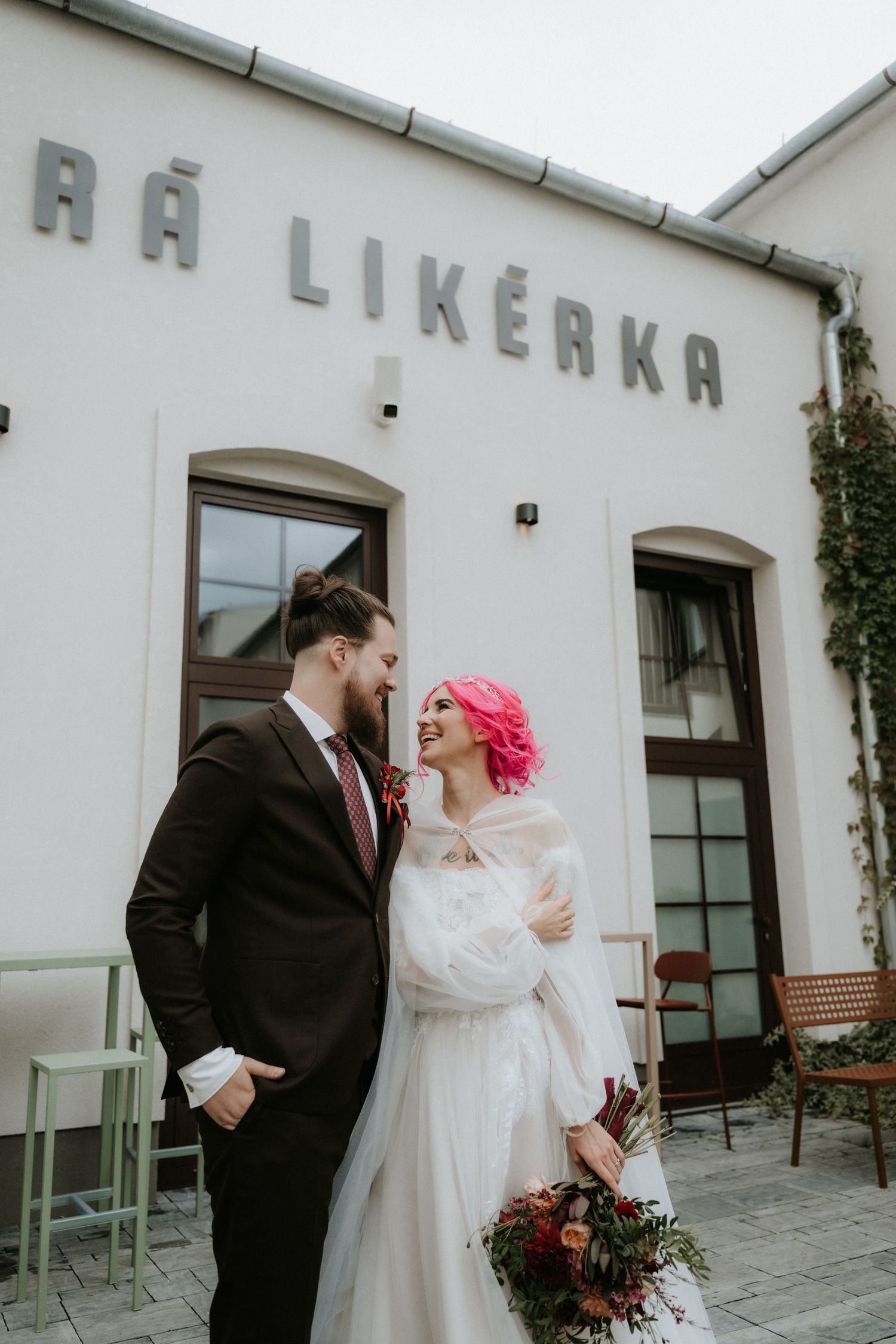 Bride with pink hair holding bouquet stands with groom in dark suit outside Stará Likérka wedding venue.