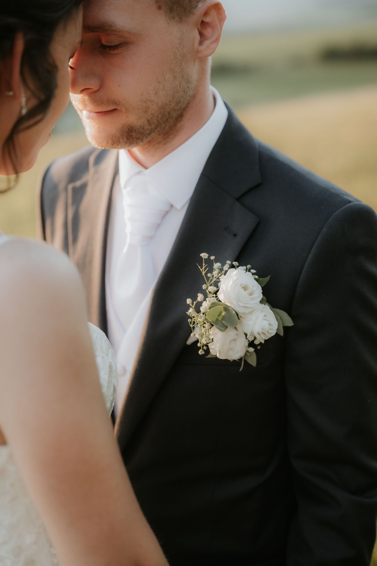 Groom in black suit with white floral boutonniere stands close to bride in soft natural light.