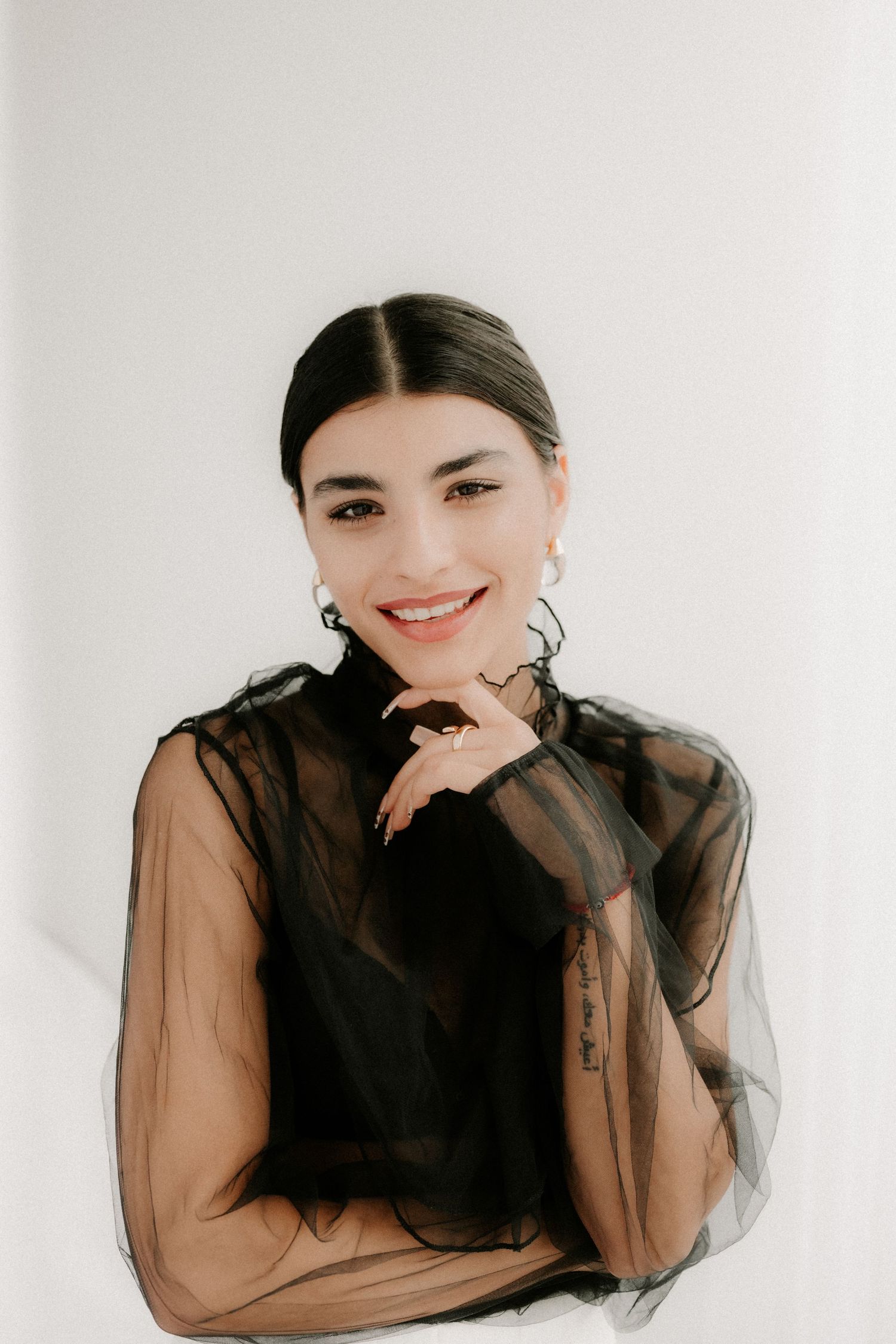 Smiling woman in black sheer blouse with hoop earrings against white background.