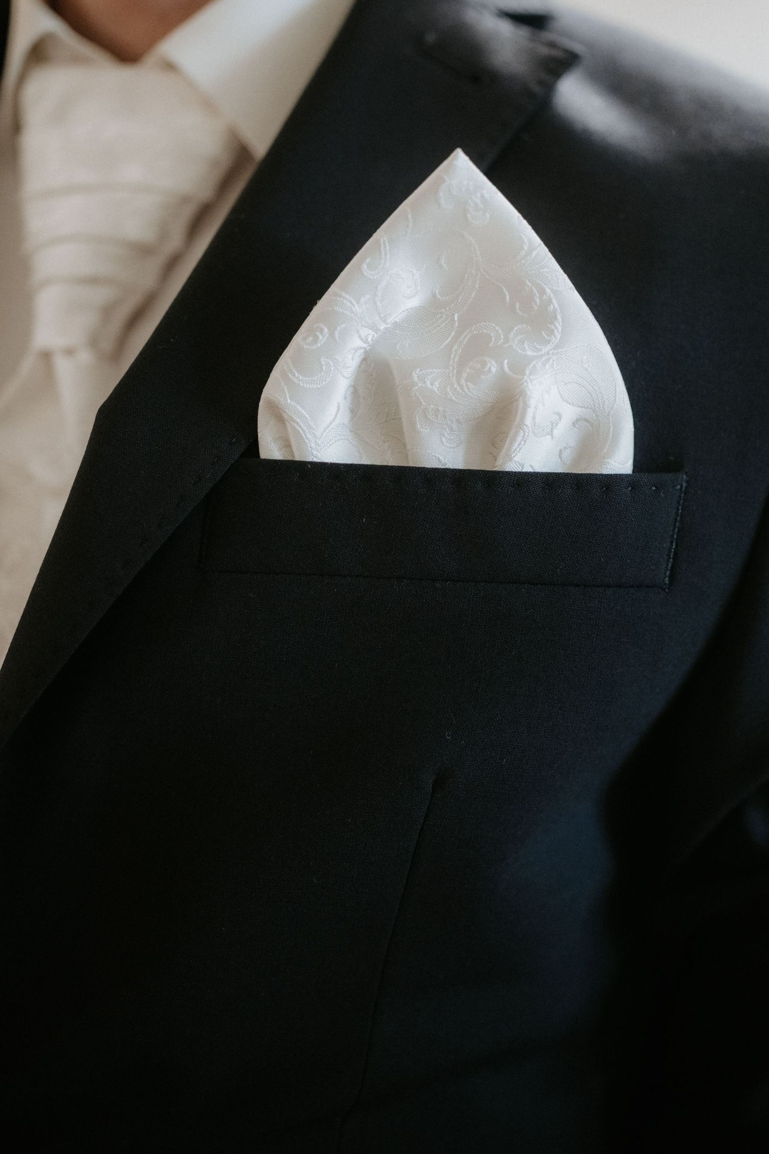 Close-up of a black tuxedo jacket with a white paisley pocket square and matching cream tie.
