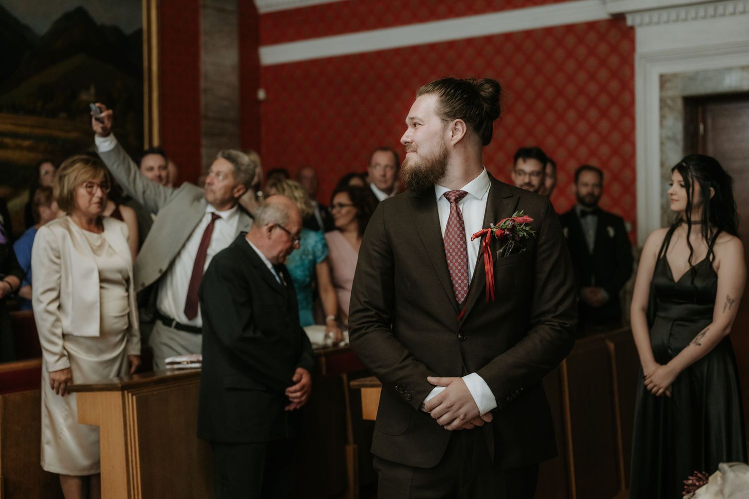 Groom in dark suit with boutonniere smiling at wedding ceremony, guests standing in elegant red-walled venue.