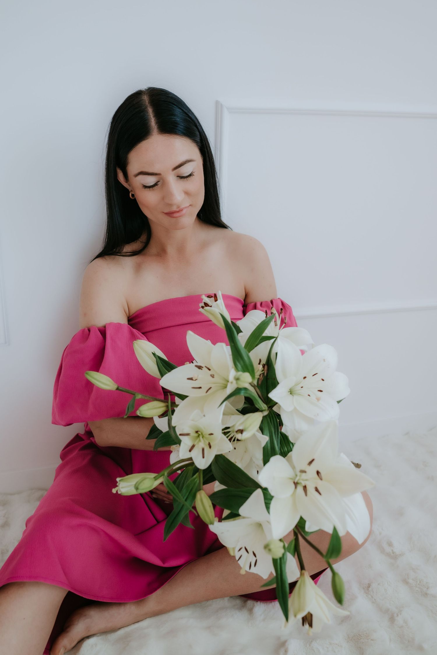 Pregnant woman in pink off-shoulder dress sitting on floor holding white lily bouquet.