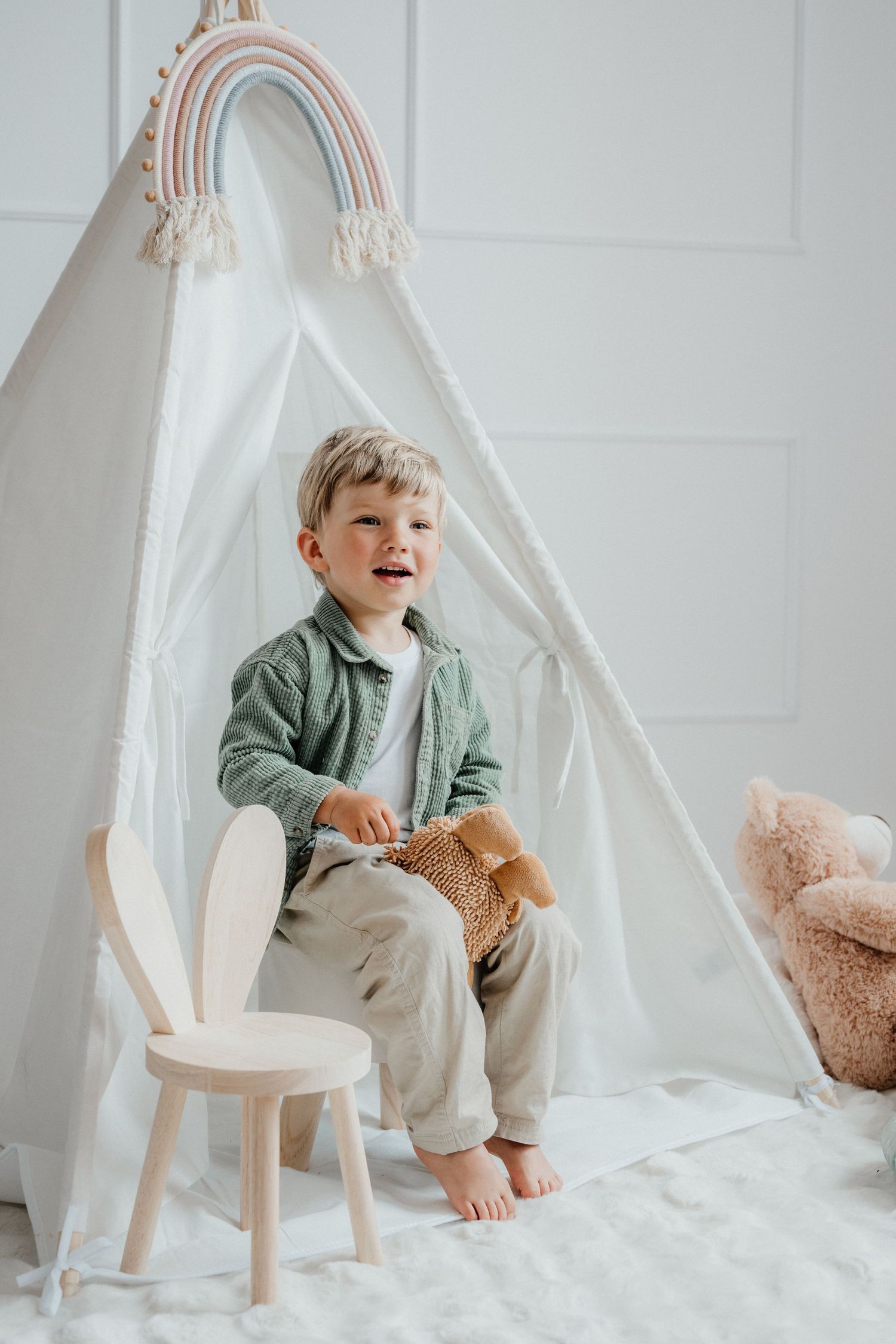 A happy laughing boy sits on a wooden bunny chair holding a toy in front of a white play teepee.