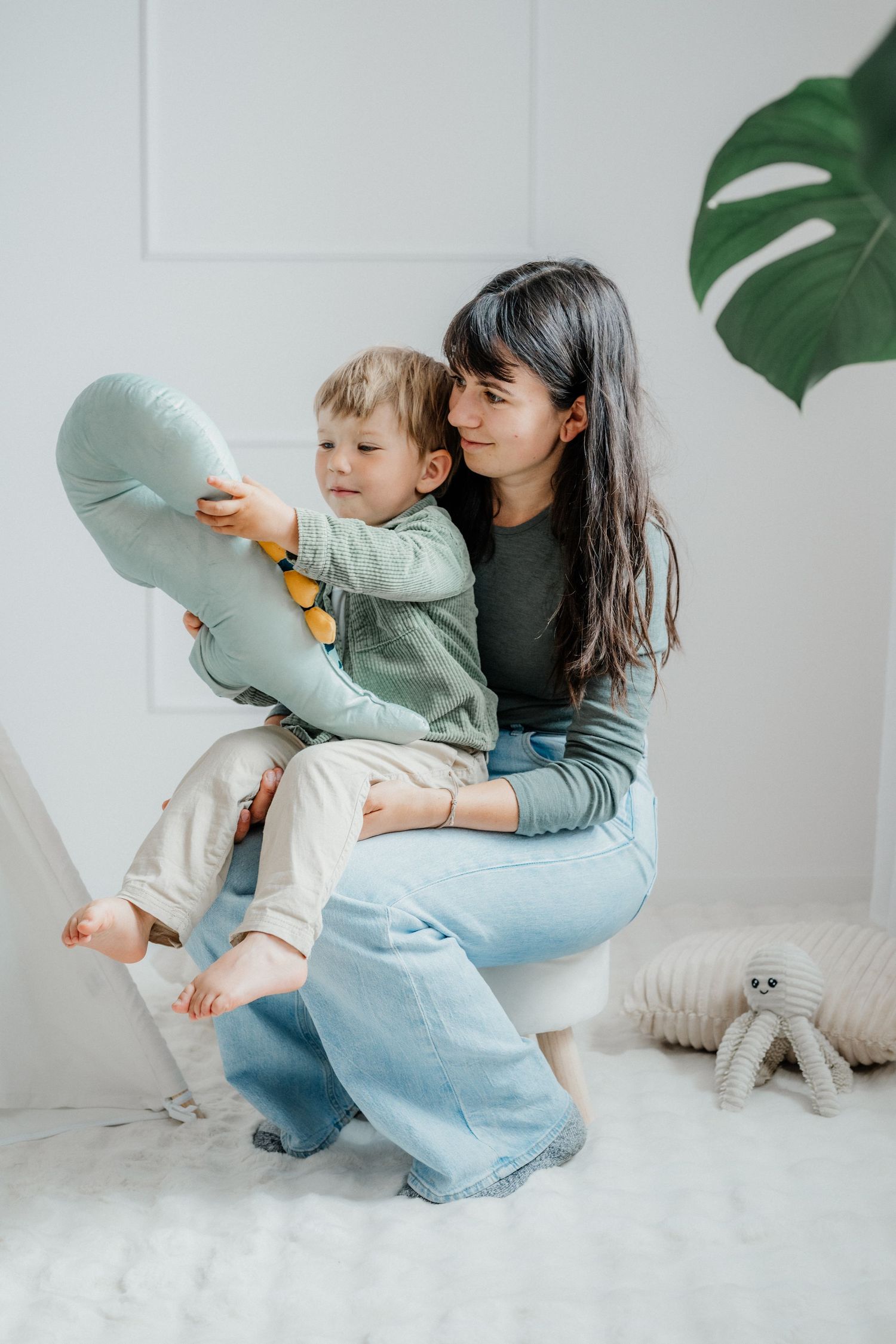 Dark-haired mother in green top and jeans holding toddler son with stuffed toy in bright white room.