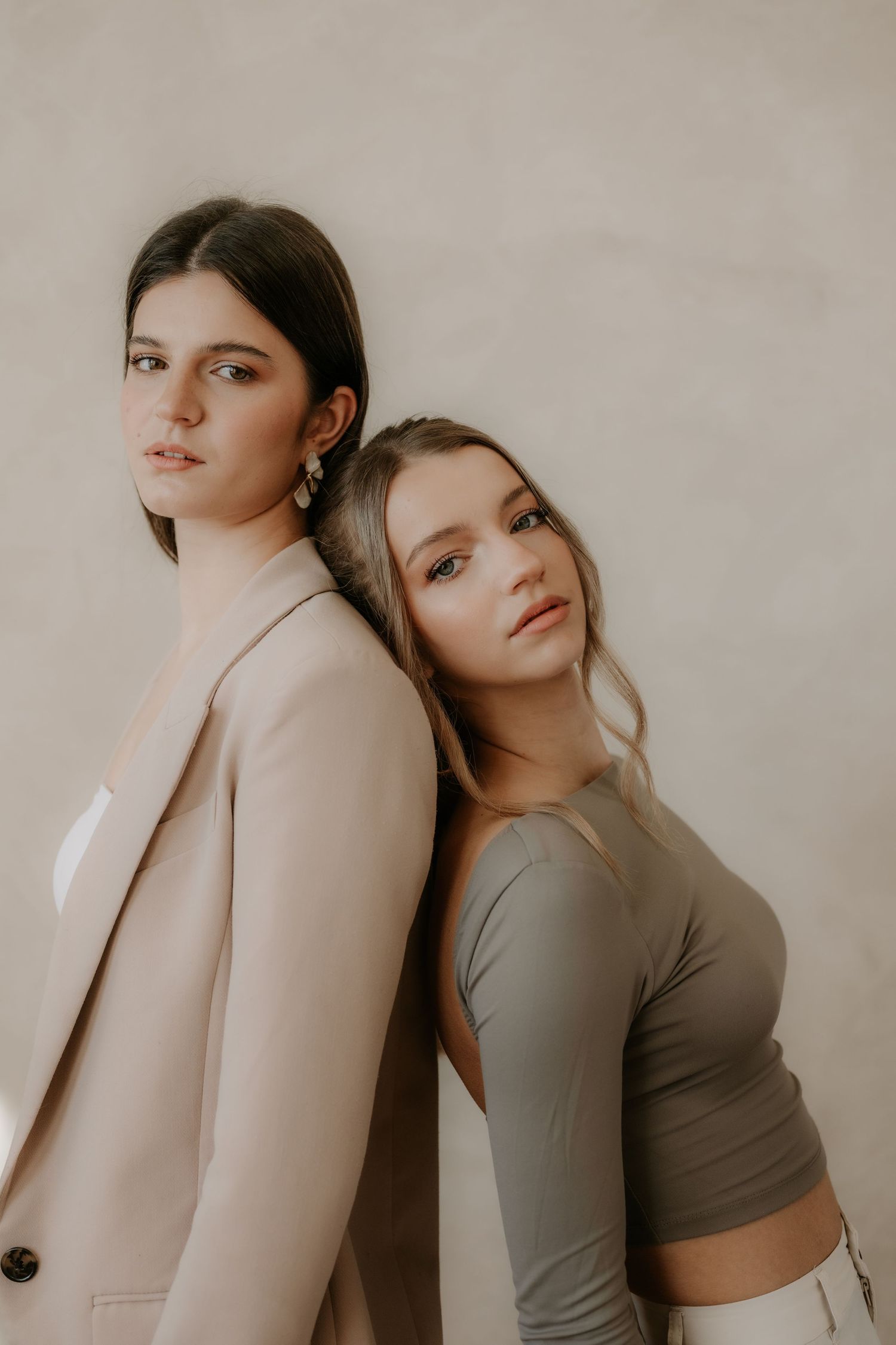 Two young women posing back-to-back against beige wall, one in blazer and one in crop top.