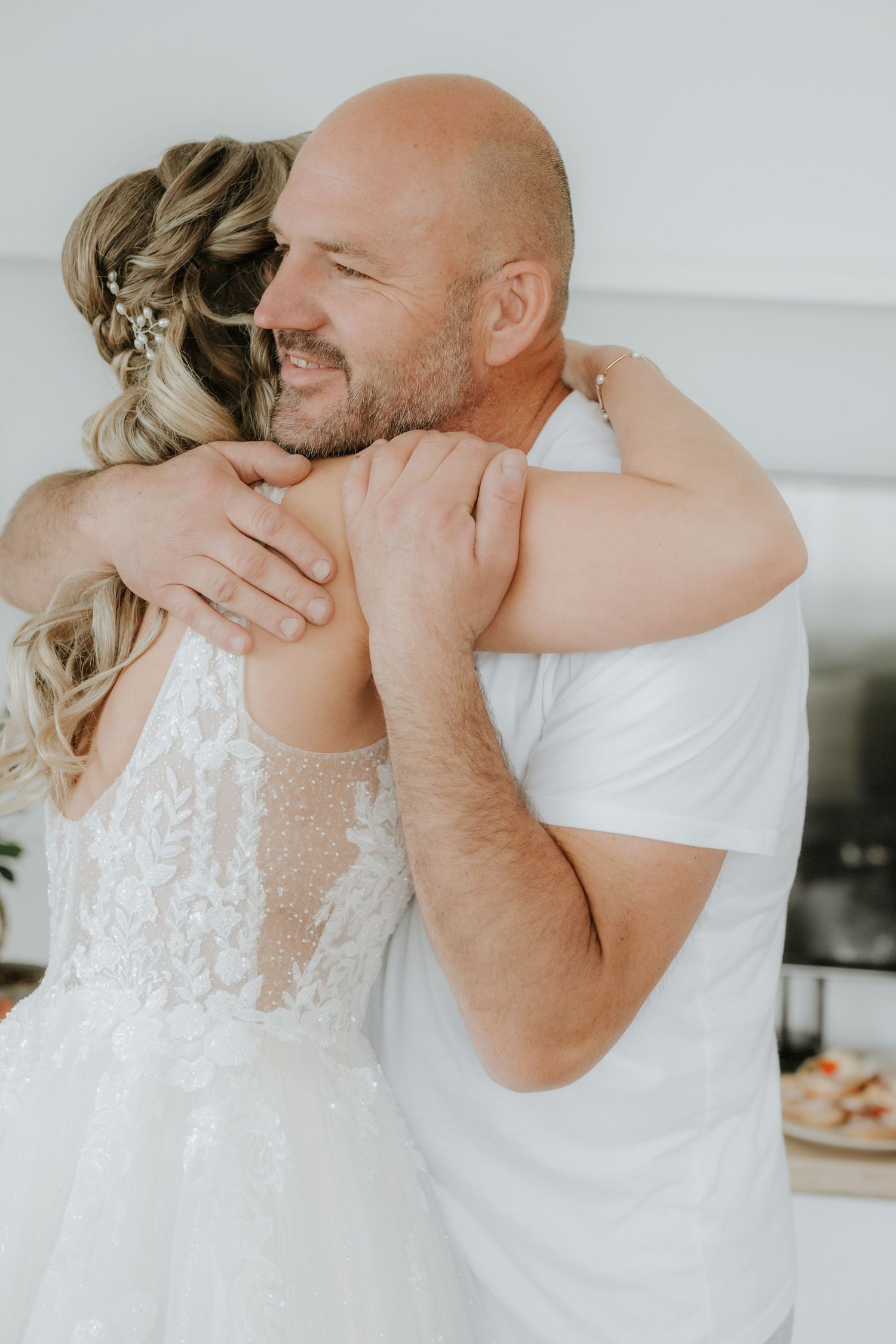 A bald groom in a white shirt embraces a bride in a lace wedding dress, both smiling warmly indoors.