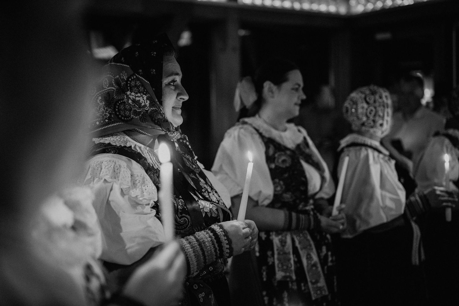 Black and white photo of women in traditional folk costumes holding candles during a religious ceremony.