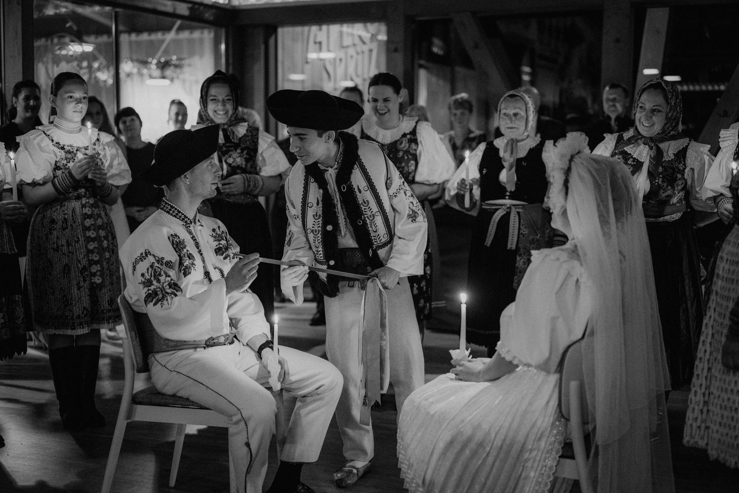 Black and white photo of people in traditional folk costumes gathered around a candlelit ceremony indoors.