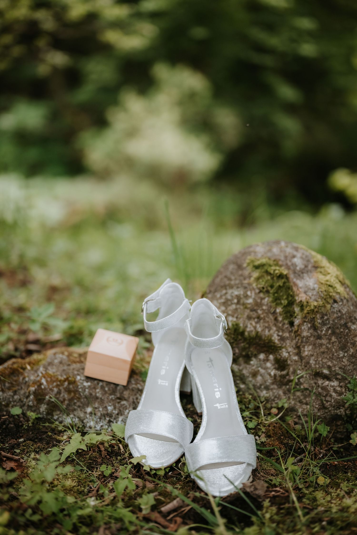 White bridal heeled sandals with ankle straps displayed on mossy rocks outdoors, with a small pink ring box nearby.