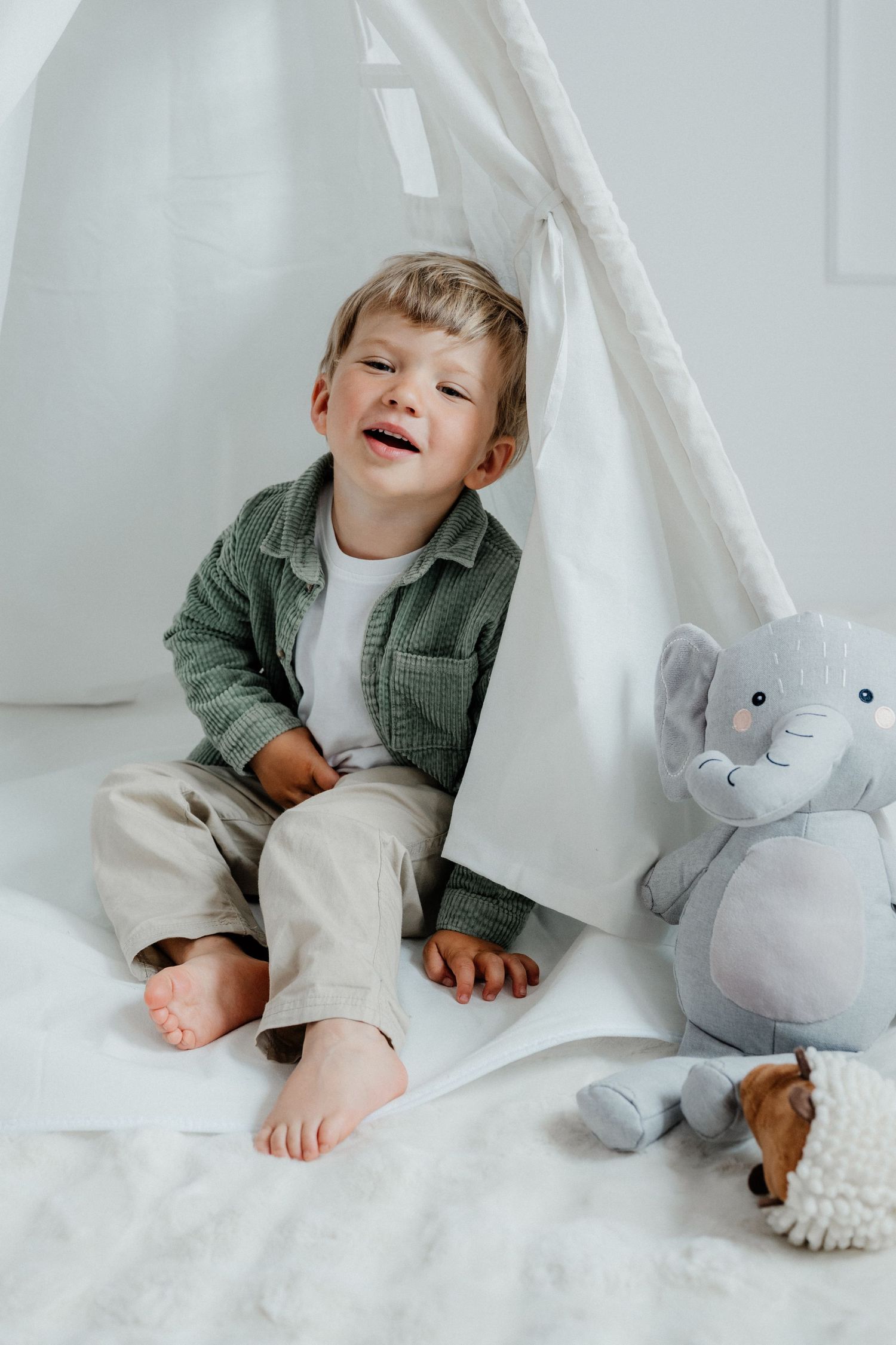 A laughing toddler boy sits in front of a white play tent with stuffed animal toys on a soft white floor.