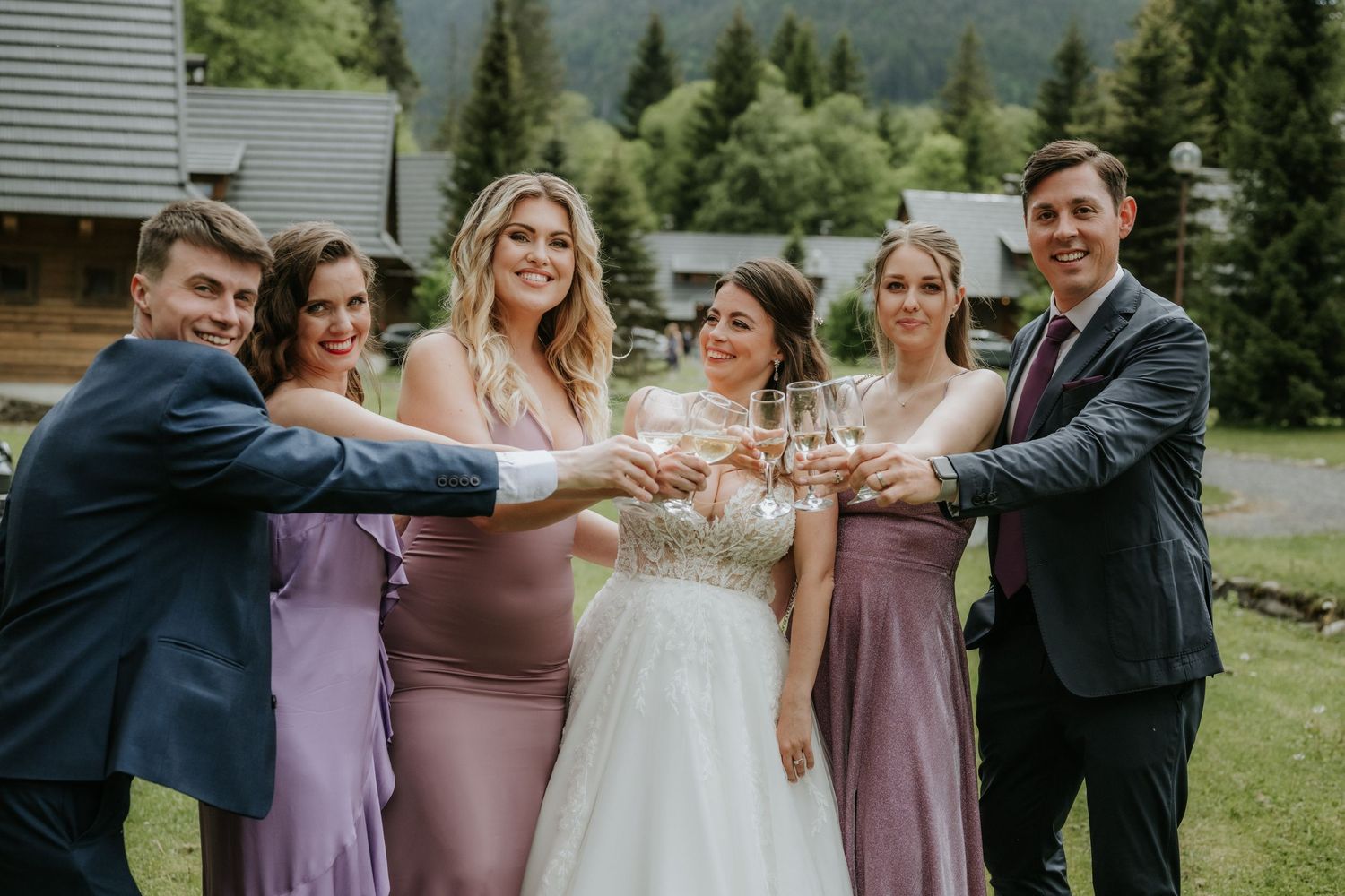 Wedding party toasting with champagne glasses outdoors, bride in white gown with bridesmaids in purple dresses and groomsmen.