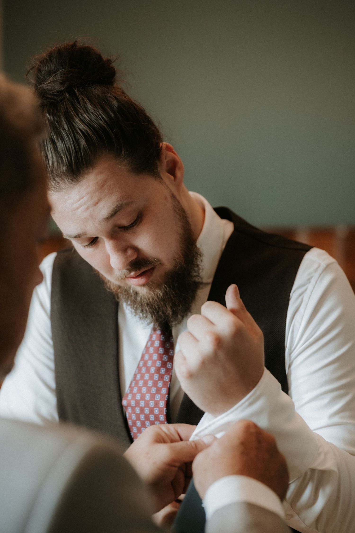 Bearded groom with man bun in white shirt, gray vest, and pink tie getting cufflinks fastened before wedding.