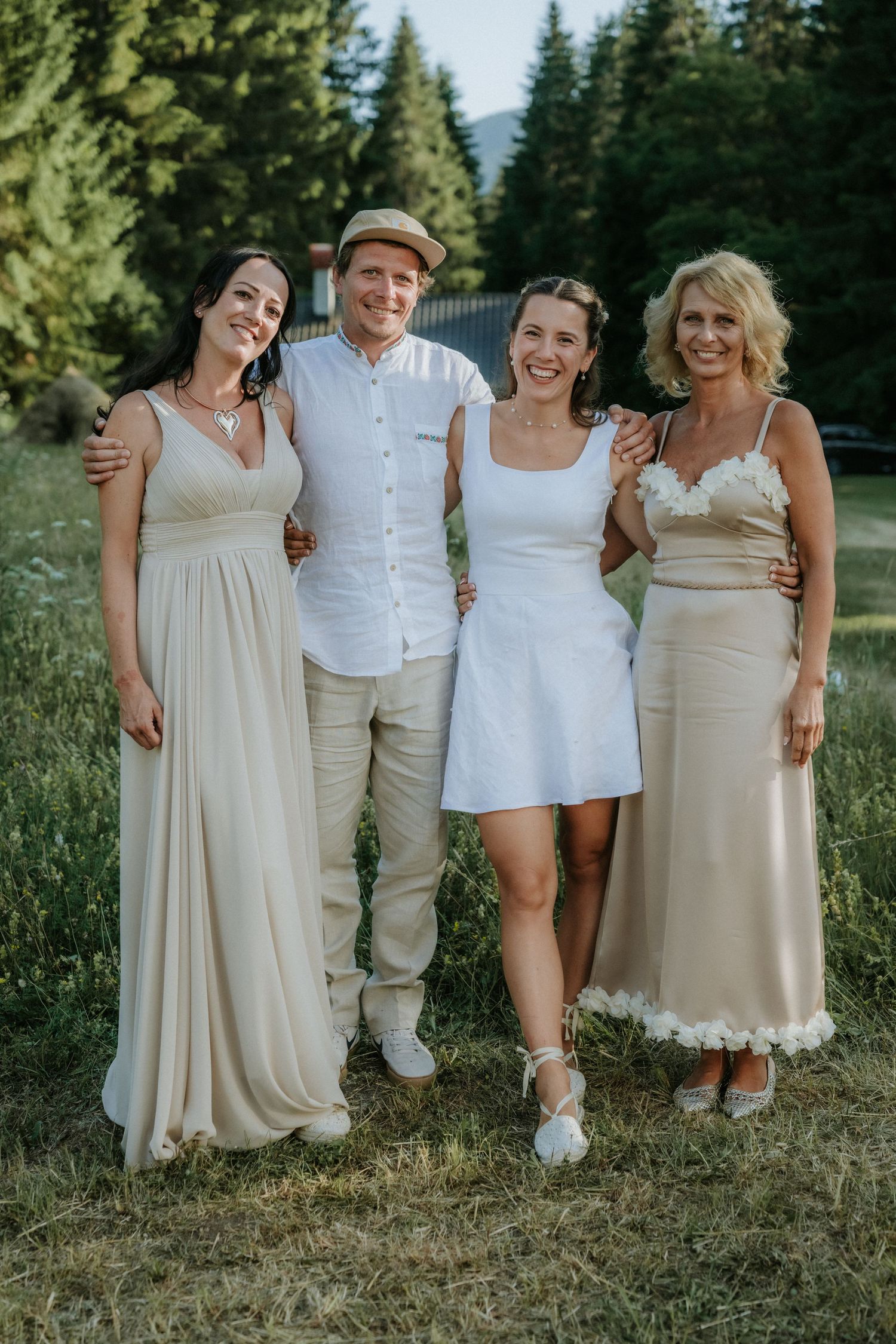 Group of four people posing outdoors at a wedding, wearing elegant white and beige outfits with trees in background.