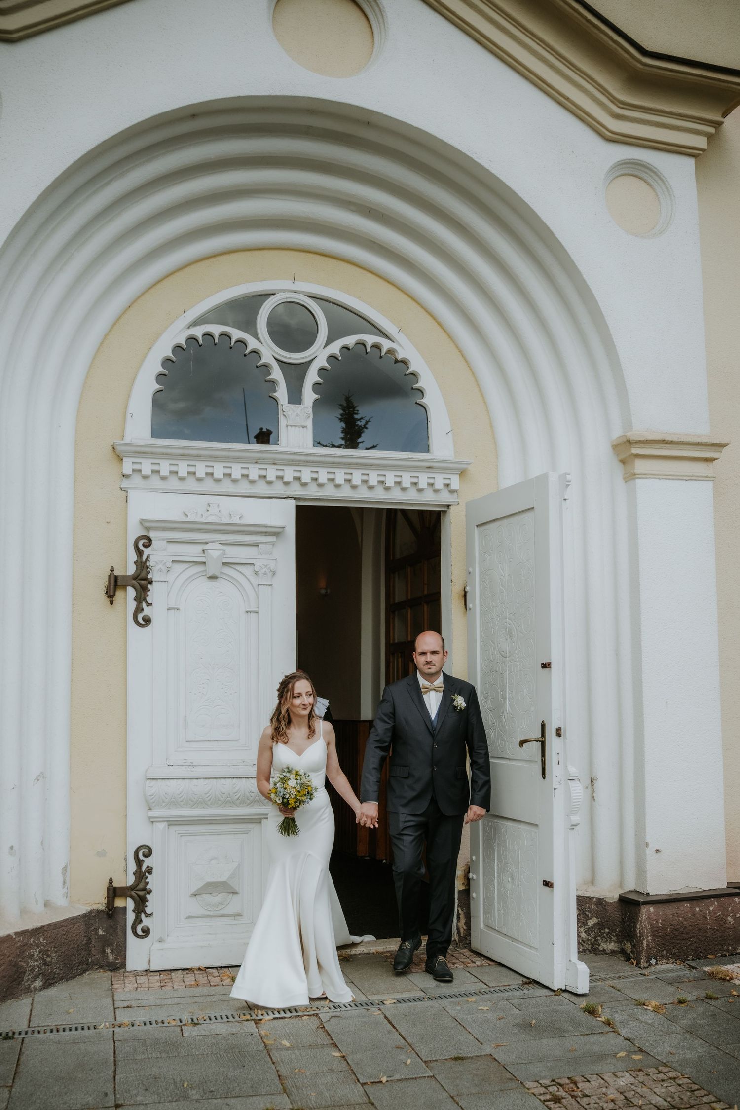 Bride and groom exit a white arched church doorway, bride holding a bouquet in a white gown.