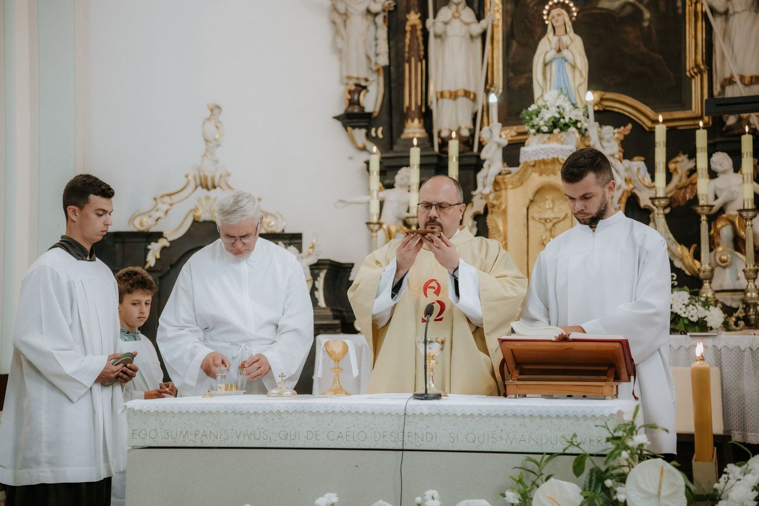 Catholic priest consecrating Eucharist at altar during Mass with altar servers in white robes in ornate church.