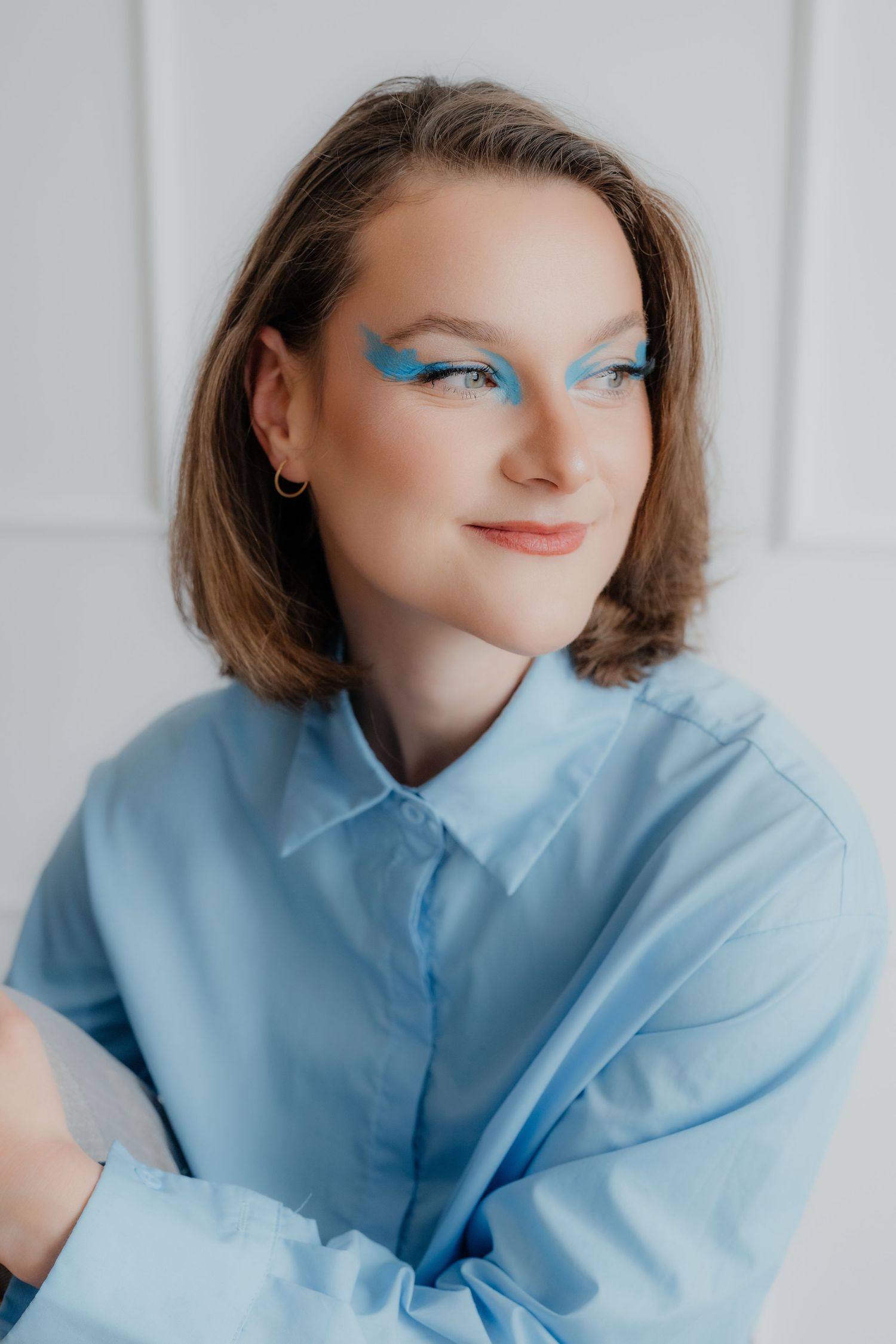 Portrait of smiling young woman with artistic blue winged eyeliner wearing blue shirt against white background.