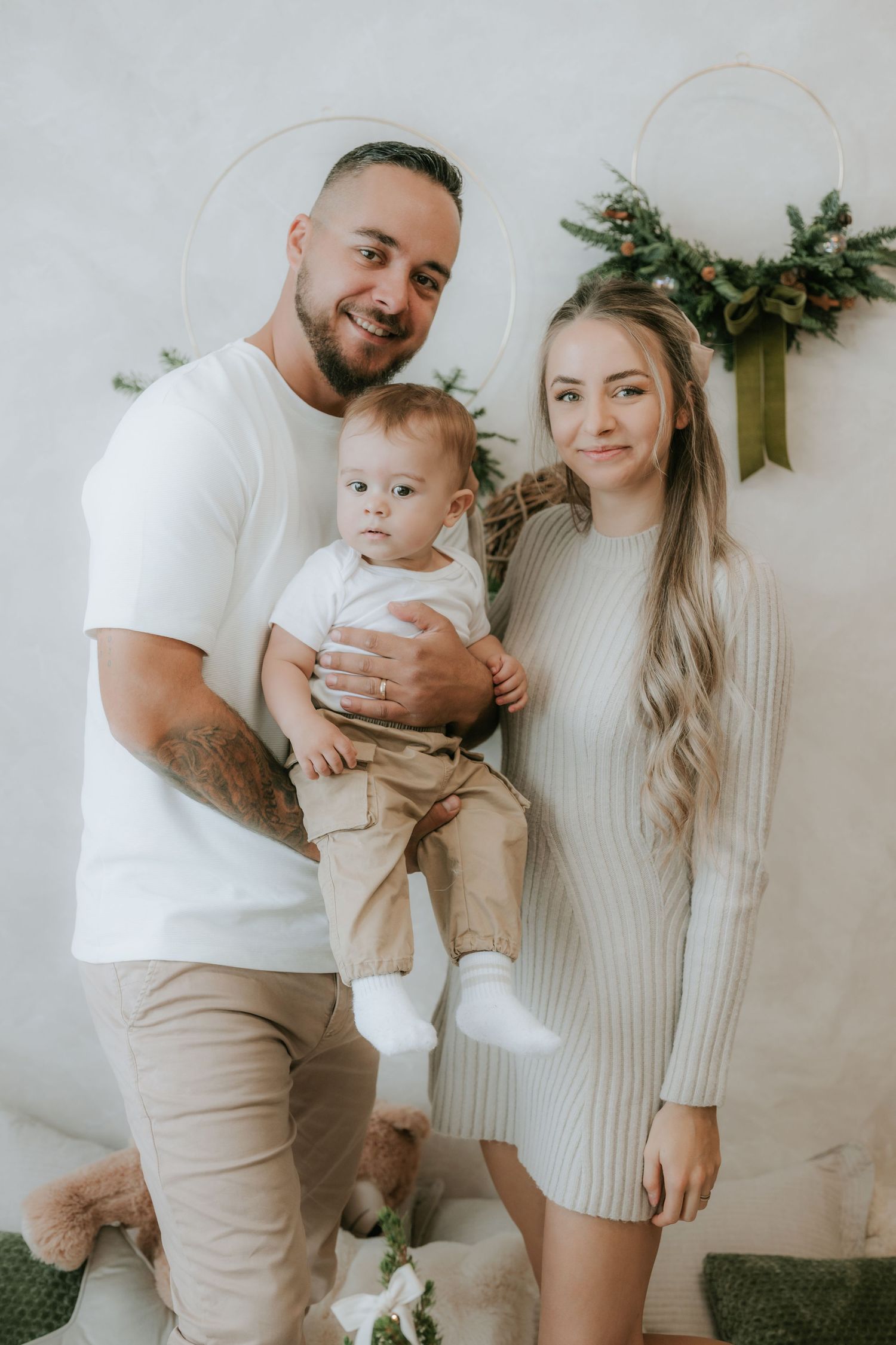 Smiling tattooed father holding baby beside blonde mother in cozy holiday decorated white room.