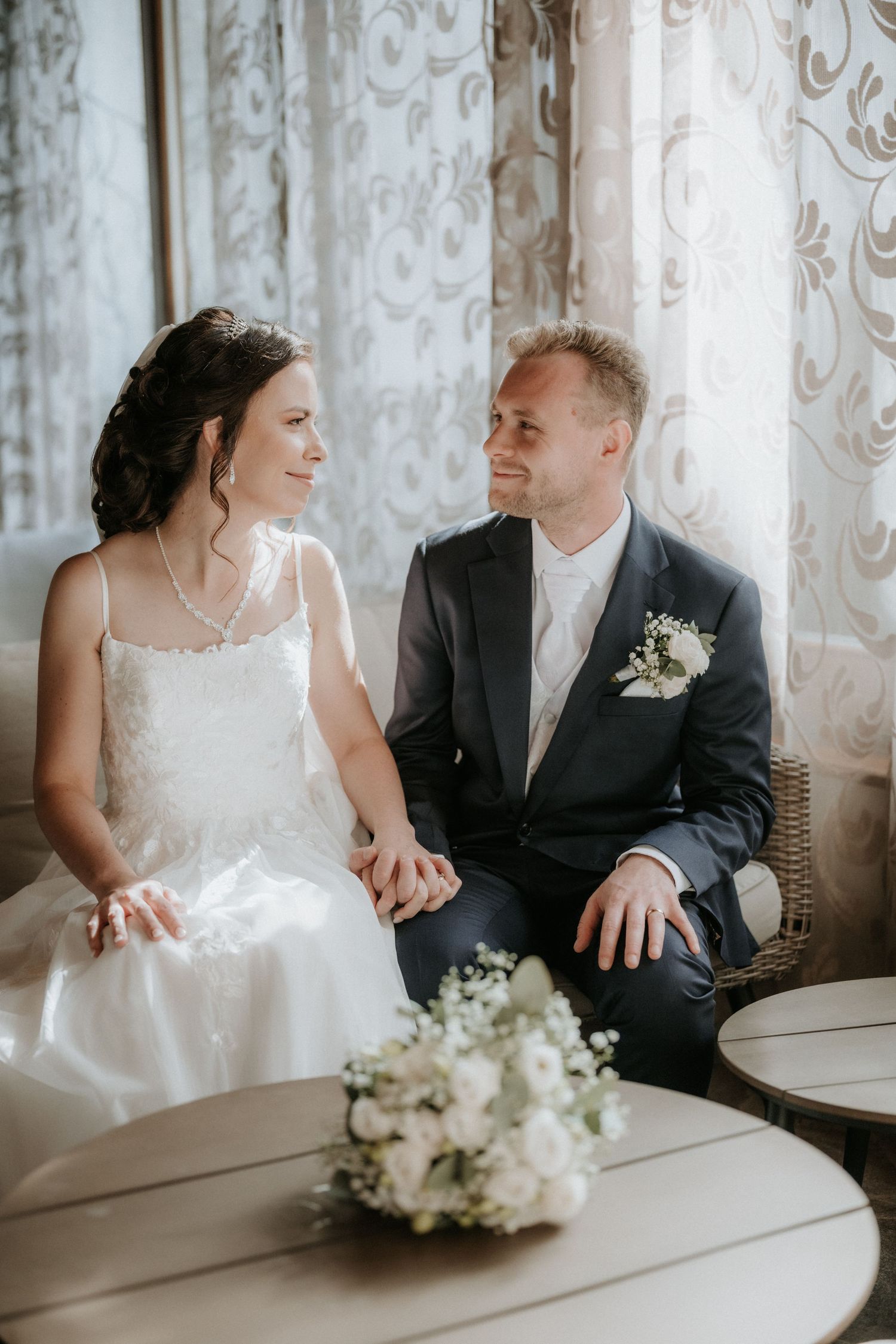 Smiling bride and groom sitting together holding hands with white floral bouquet on table.