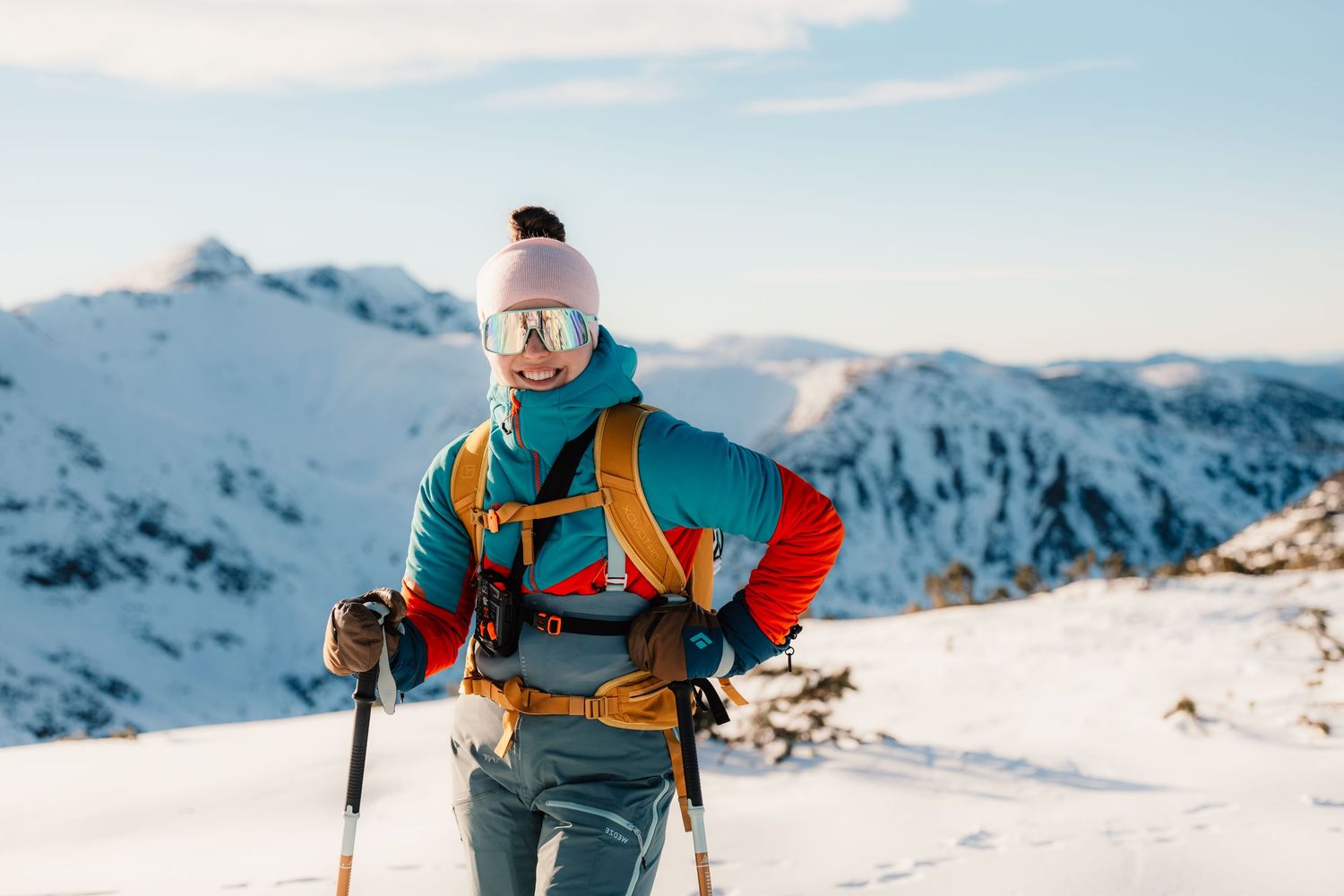 Close-up of a backcountry skier with orange backpack and goggles trekking through deep snow in winter mountains.