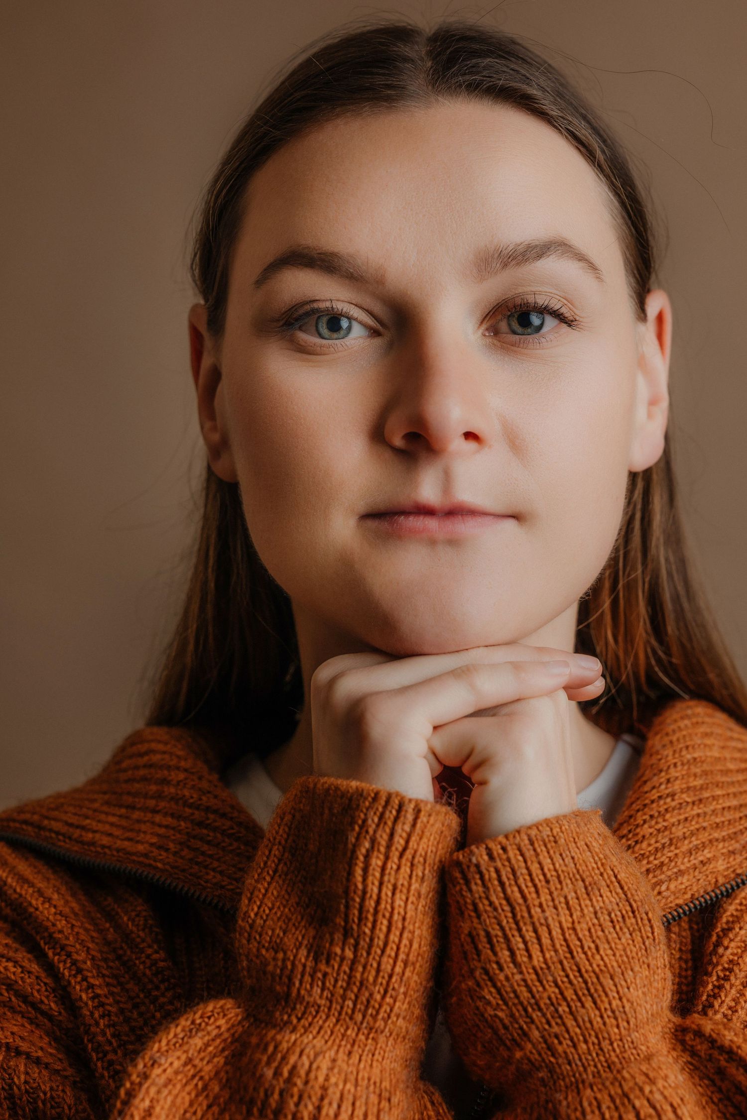 Young woman with blue eyes in rust orange knit sweater resting chin on hands against beige background.