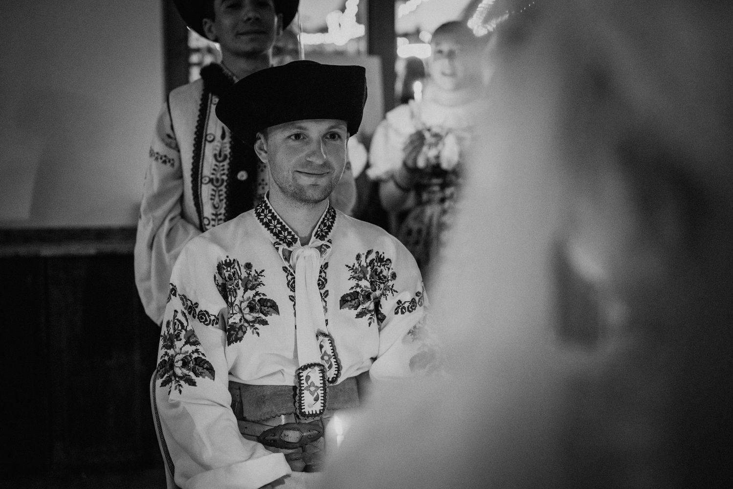 Man in traditional embroidered folk costume and black hat holding a candle at a ceremony, black and white photo.