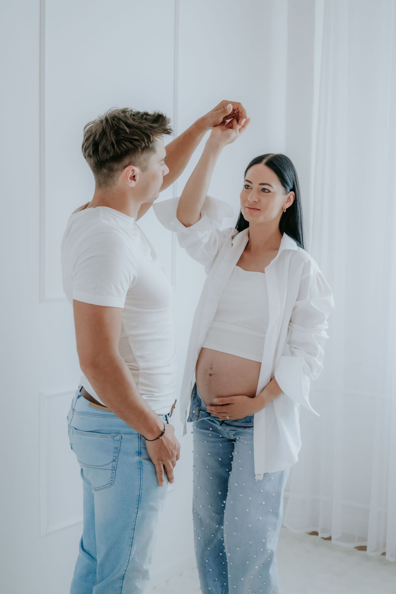 Pregnant couple dancing together in a bright white studio, woman cradling her baby bump.